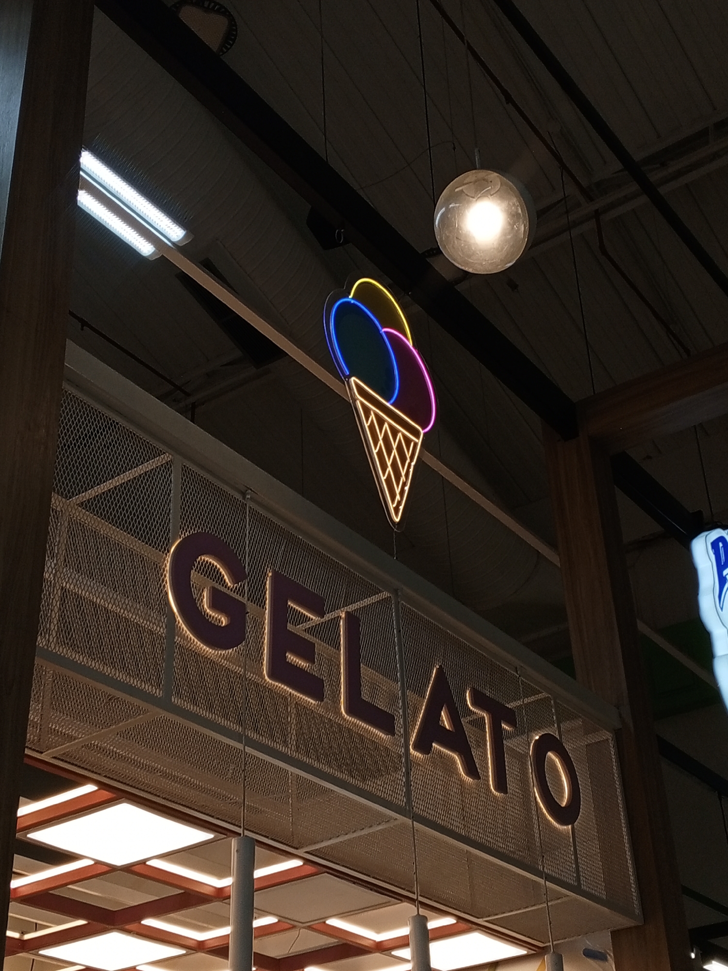 The image shows an interior view of a structure with a "GELATO" sign in large, illuminated letters. The sign is mounted on a mesh framework above a well-lit lower level. Above the sign, there is an illuminated ice cream cone graphic in neon colors. A spherical pendant light is suspended from the ceiling above the scene. The image does not contain any information pertaining to the city of Porlamar, Venezuela or any specific location.