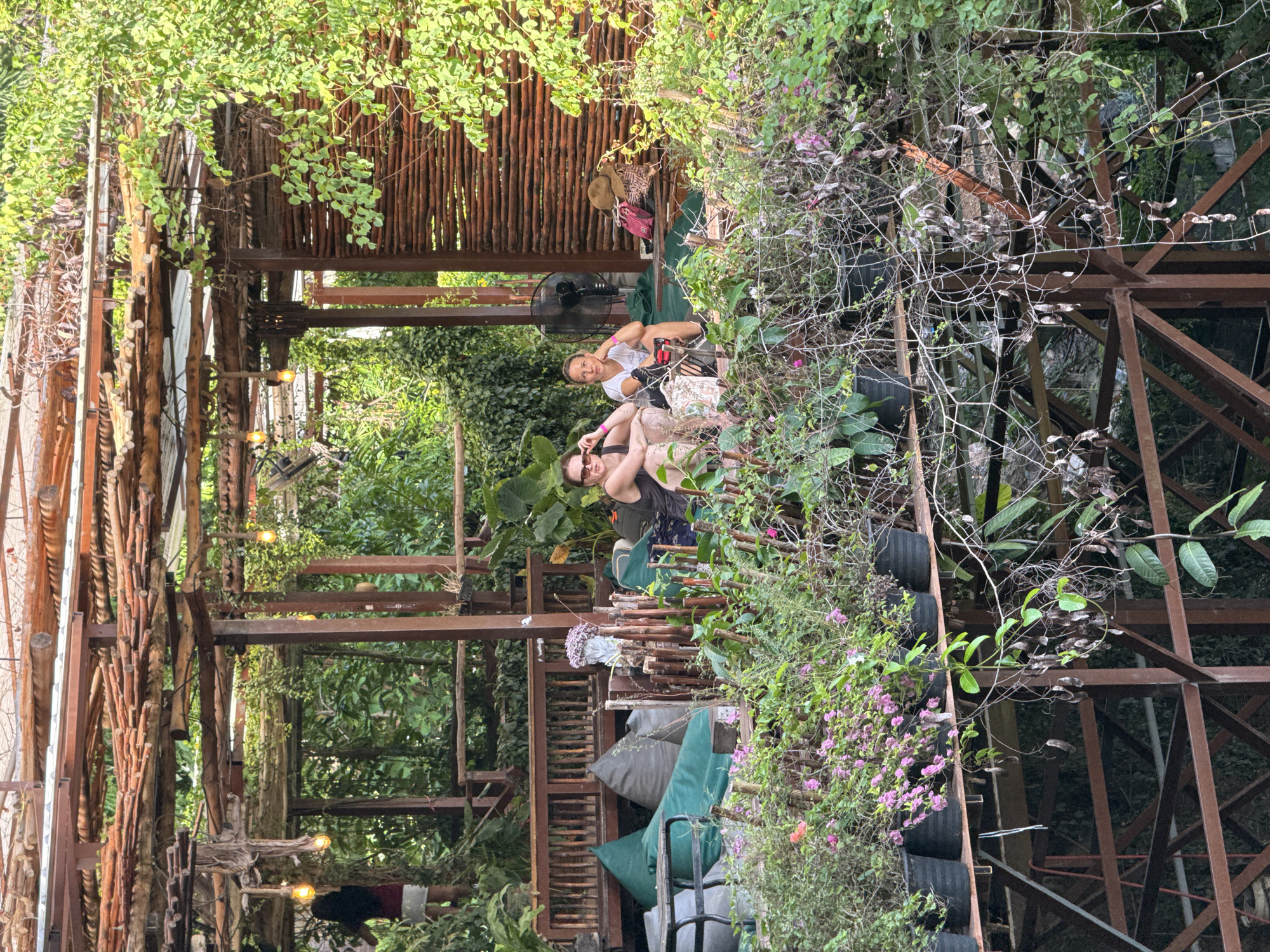 Two women are seated on a balcony amidst lush vegetation. The woman on the left is wearing sunglasses and a dark top, with her legs crossed. The woman on the right is wearing a white top and has her hand on her chin. Behind them, a straw hat and some bags are placed on a bench, next to a standing fan. The balcony is part of an open-air structure with a rustic wooden roof and walls made of woven branches. A variety of potted plants, including those with pink flowers, are arranged on a railing in the foreground. The background is filled with dense green foliage. The scene suggests an outdoor seating area, possibly a cafe or a lounge, in a tropical environment, consistent with the Ko Samui District, Thailand.