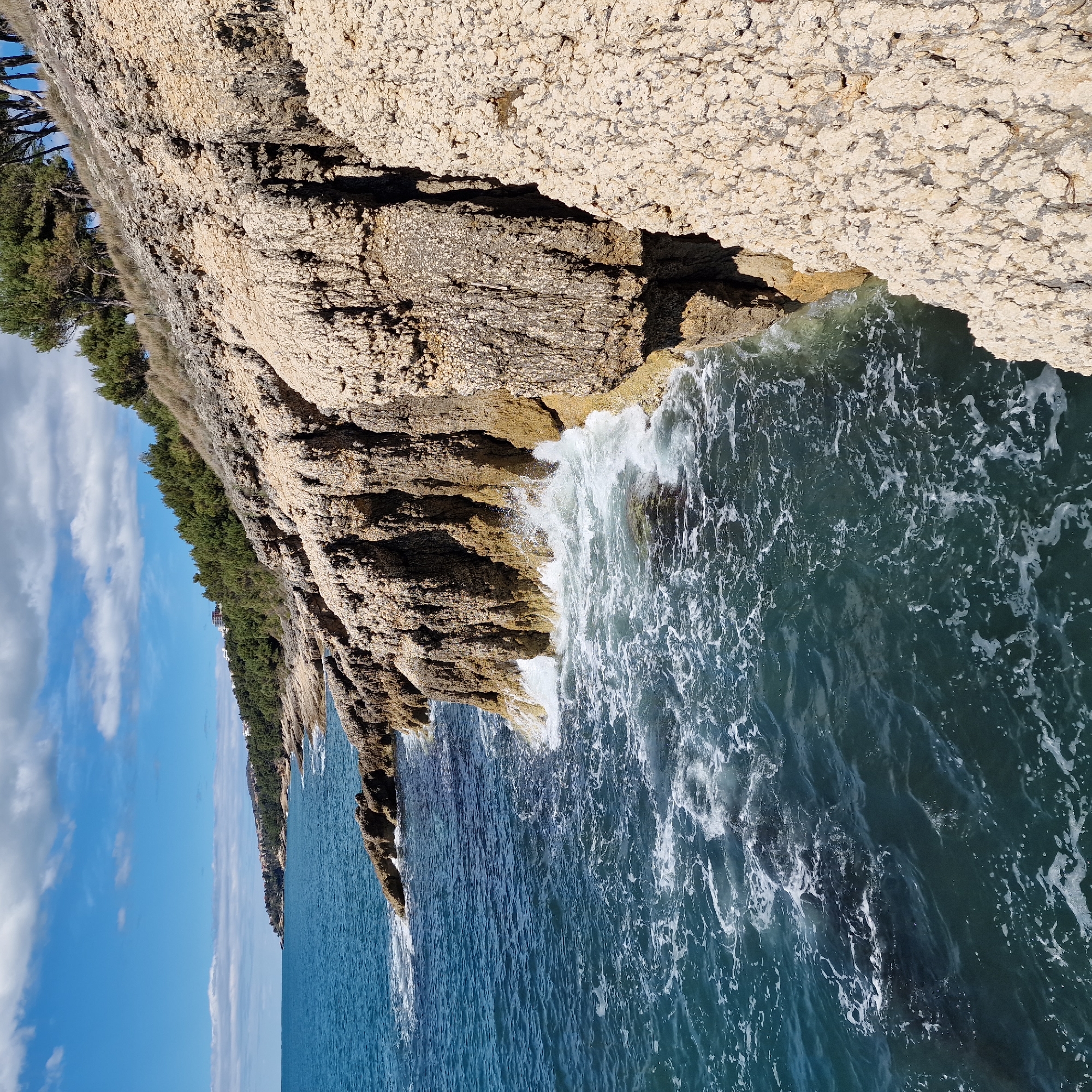 The image shows a rocky coastline along the Adriatic Sea in Ulcinj, Montenegro. The scene depicts tall, jagged cliffs with sparse vegetation at the top meeting the sea. Waves crash against the base of the cliffs, creating white foam. The water transitions from a lighter turquoise near the shore to a deeper blue further out. The sky is blue with scattered white clouds.
