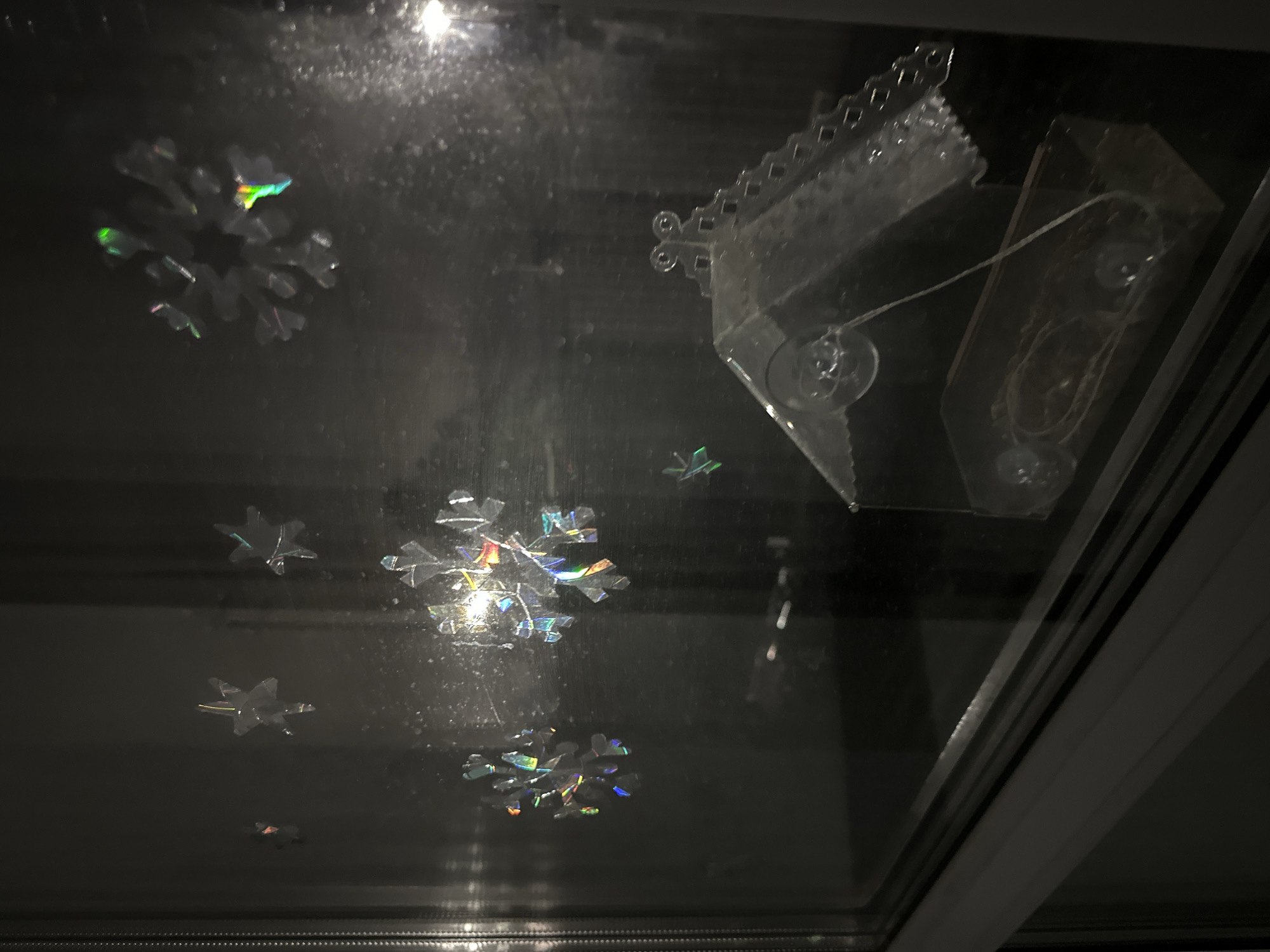 The image captures a close-up, upward-angled view of a dark, reflective surface, likely a window or glass panel, adorned with translucent decorations. Several snowflake-shaped ornaments, made of a clear or iridescent material, are adhered to the surface. These snowflakes exhibit a colorful, holographic shimmer where light reflects off them, creating small rainbow effects. In the upper right portion of the frame, a larger, more complex clear plastic decoration is visible. This object resembles the outline of a house or building, featuring decorative scalloped edges along its roofline. It is attached to the glass using two visible suction cups. A thin, light-colored string or wire extends from this house-shaped ornament to another smaller, clear rectangular plastic piece, also secured to the glass with suction cups. The background seen through the glass is uniformly dark, suggesting either nighttime outside or a dimly lit interior space. No people or discernible activity are present. The scene is static, focusing solely on the decorative items. There is no visible text.