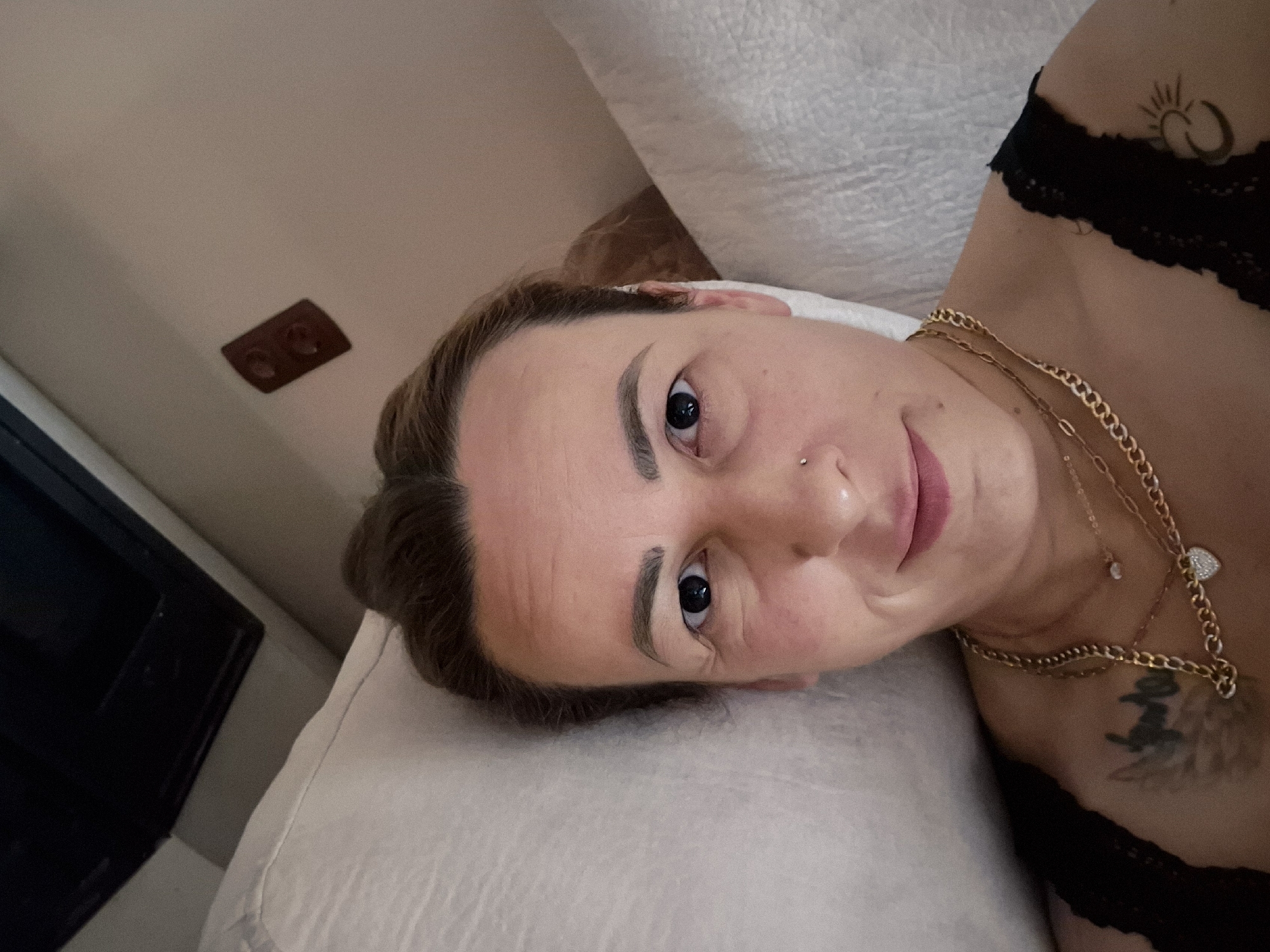 A selfie is taken from a low angle, capturing a person lying on a bed, propped up against white pillows. The person is looking directly at the camera with dark eyes and a slight smile. They have their hair pulled back and are wearing a black top adorned with multiple gold necklaces, one with a heart-shaped pendant. A nose ring is visible on their right nostril. Tattoos are present on the left shoulder area, and another tattoo of a crescent moon and sun symbol is visible on the right shoulder. The background shows a dark rectangular object above the pillows, possibly a television screen, and a brown electrical outlet on the wall. The lighting is soft, suggesting an indoor setting.