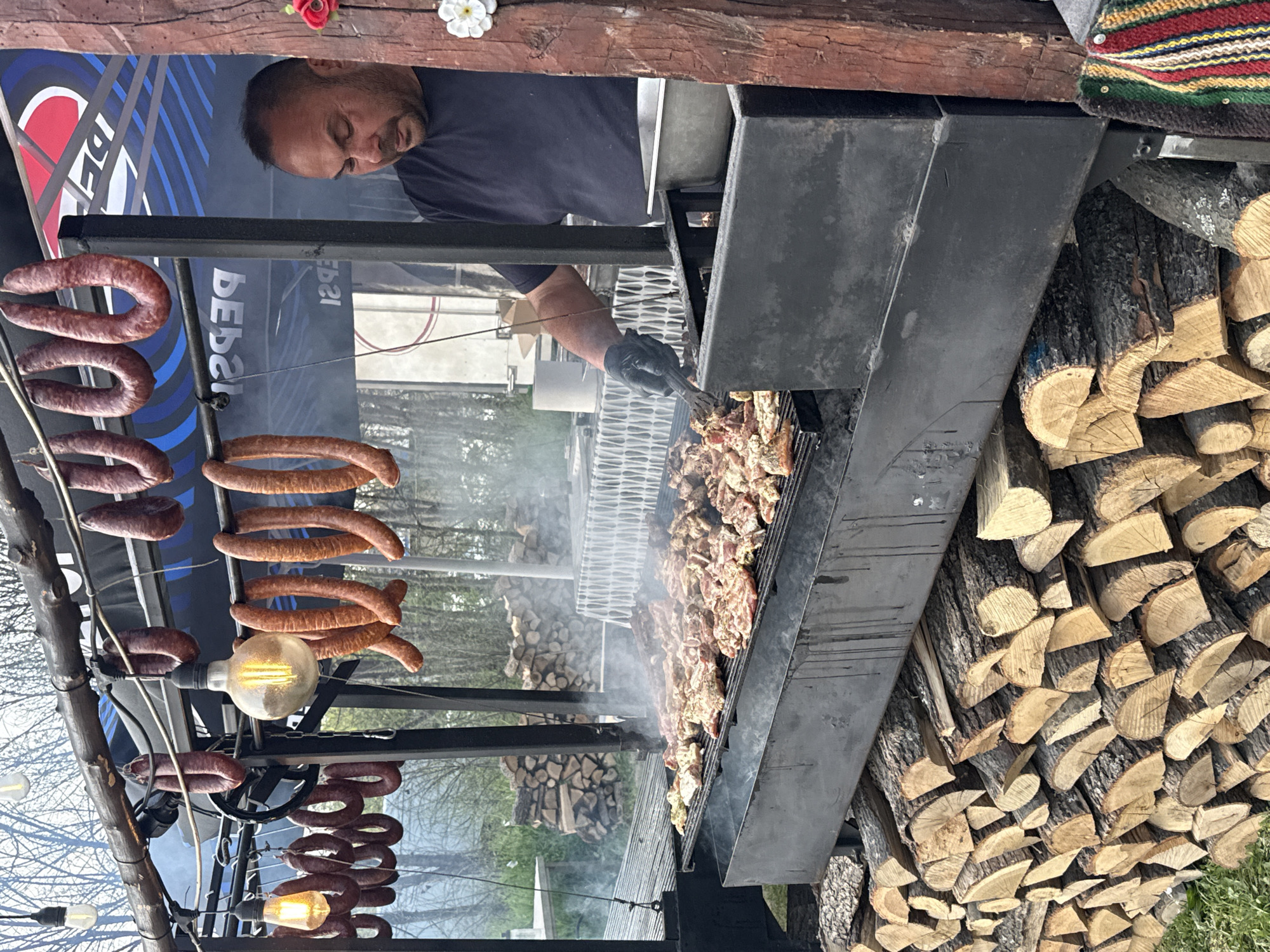 The image captures an outdoor grilling scene, possibly at a restaurant or food stall in Plovdiv, Bulgaria. A man is seen tending to a large grill filled with pieces of meat. Smoke rises from the grill, indicating active cooking. To the left, strings of sausages hang above the grill, illuminated by decorative light bulbs. A stack of firewood sits beside the grill, suggesting a wood-burning heat source. In the background, a Pepsi advertisement is visible. The environment appears to be a semi-urban outdoor setting, possibly a park or public space.