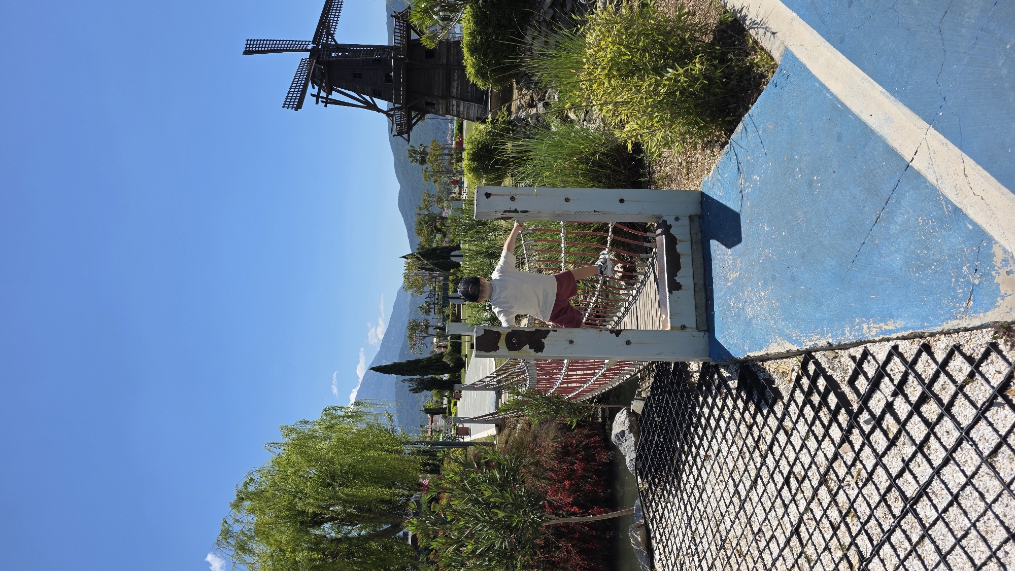 The image depicts a child positioned on a rope bridge within a park or recreational area in Fethiye, Turkey. The child is wearing a white shirt and maroon shorts. The bridge has weathered metal railings and spans over an area containing vegetation. In the background, a traditional windmill is visible against a backdrop of mountains and a clear blue sky. To the right of the bridge is a blue painted surface with a white stripe. Below the bridge, there is a black plastic grid containing white stones.
