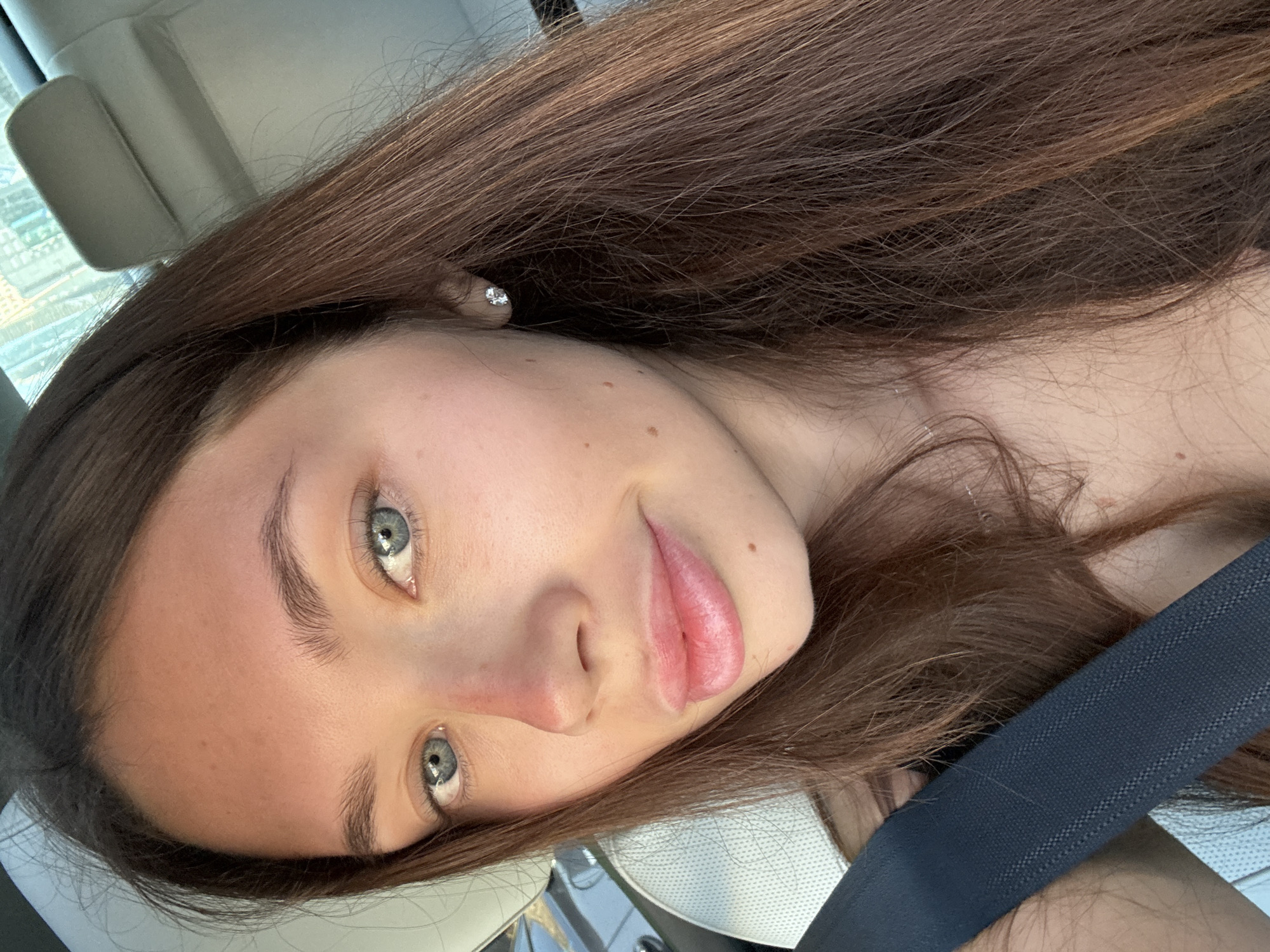 A close-up, tilted selfie of a young woman with light skin, grey-blue eyes, and brown hair. She is wearing a seatbelt in what appears to be a car. The interior of the car features light-colored leather seats. The background is blurry and indistinct, and there is no information to confirm that the location is Ramat Gan, Israel. No specific location can be determined from this image.