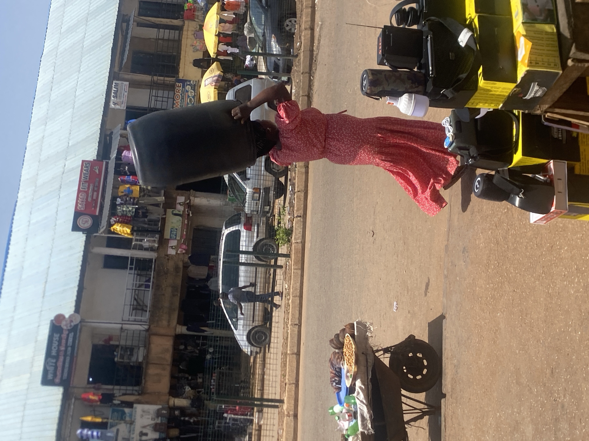 A woman in a red floral dress carries a large black container on her head, walking along a paved street. She appears to be navigating a busy marketplace with buildings and stalls lining the road. Various items, including boxes, electronics, and umbrellas, are visible on stalls. A white van is parked on the side of the road, and other people can be seen in the background, suggesting a lively daytime scene. Signs on the buildings indicate businesses, with one clearly reading "SALIM" and another "MORE". There is also a sign for "WHITE HOUSE". The overall atmosphere seems to be one of commerce and daily activity in an outdoor market setting. The lighting suggests it is daytime, likely during fair weather.