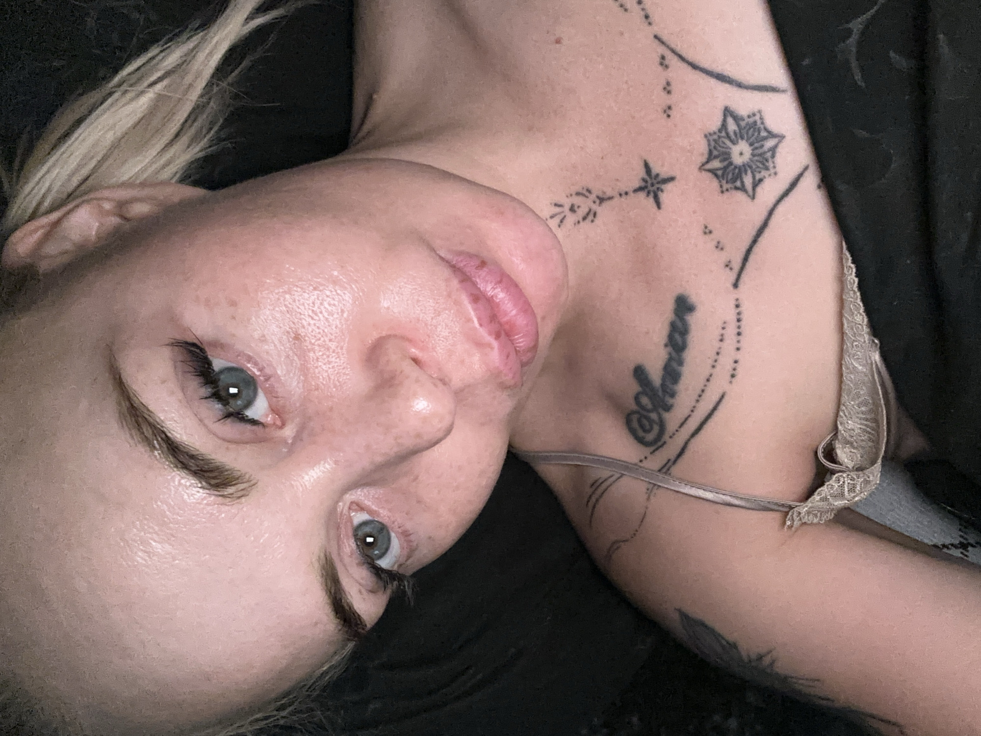 A close-up, high-angle, indoor, eye-level, natural-light shot shows a woman with blonde hair pulled back, looking towards the camera. She is wearing a beige, lacy camisole. She has light blue eyes, fair skin with freckles, and full lips. She has a tattoo on her chest with the word "Amor" and a floral design with a star. The background is dark and out of focus.