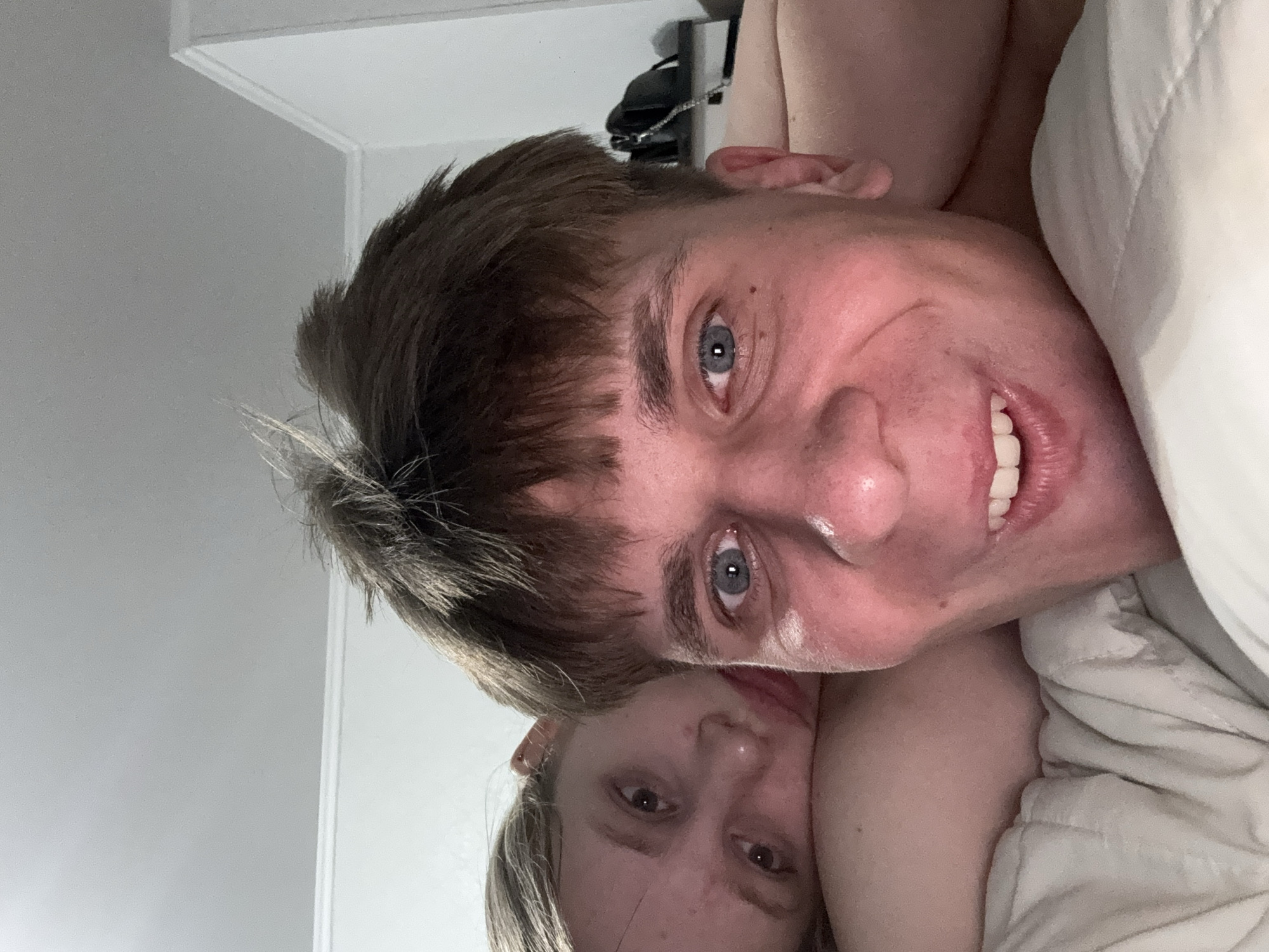 The image is a close-up, low-angle selfie of a young Caucasian man and woman in bed. The man is in the foreground, looking directly at the camera with a wide smile. He has short, messy brown hair and blue eyes. The woman is behind him, resting her head on his shoulder. She has long blonde hair and is looking slightly off-camera with a neutral expression. They appear to be in a bedroom, with a white wall and ceiling visible in the background. The image does not provide any specific details about the location in Tiraspol, Moldova.