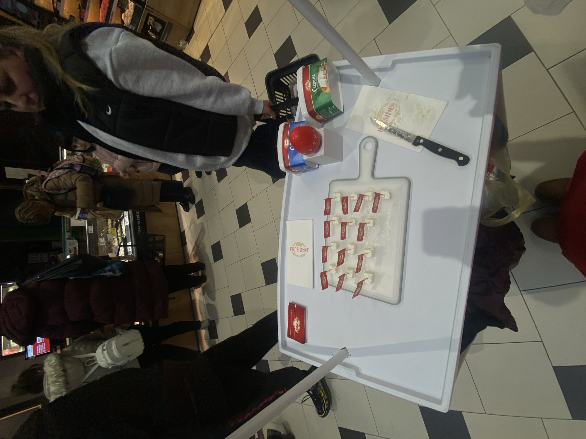 The image shows a promotional display for President brand cheese in what appears to be a supermarket setting, likely located in Sofia, Bulgaria, given the Cyrillic text on the packaging.
The display consists of a white table covered with a white tray, a tray with President cheese samples, and a small chopping board with more cheese samples with flags attached. Adjacent to the cutting board is a knife. Also visible are a folded President napkin, a stack of President pamphlets, cheese and red pepper sample jars in a basket on the top of the tray.
Several individuals are present around the display, including at least one person reaching into a basket. Other customers are passing by in the background. The scene is indoors, on a tiled floor. The lighting is somewhat dim.