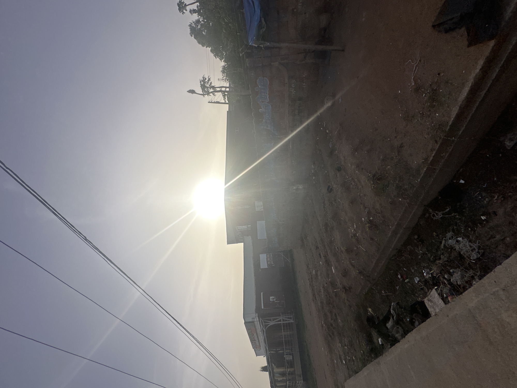 The image shows a street scene in Bukuru, Nigeria. The sun is prominent in the sky, creating glare. In the foreground, concrete and trash-strewn earth are visible. A building with signage appears along the right side, potentially a liquor store. Power lines stretch across the upper left. There is graffiti on the wall. Vegetation and a few palm trees are visible in the background.