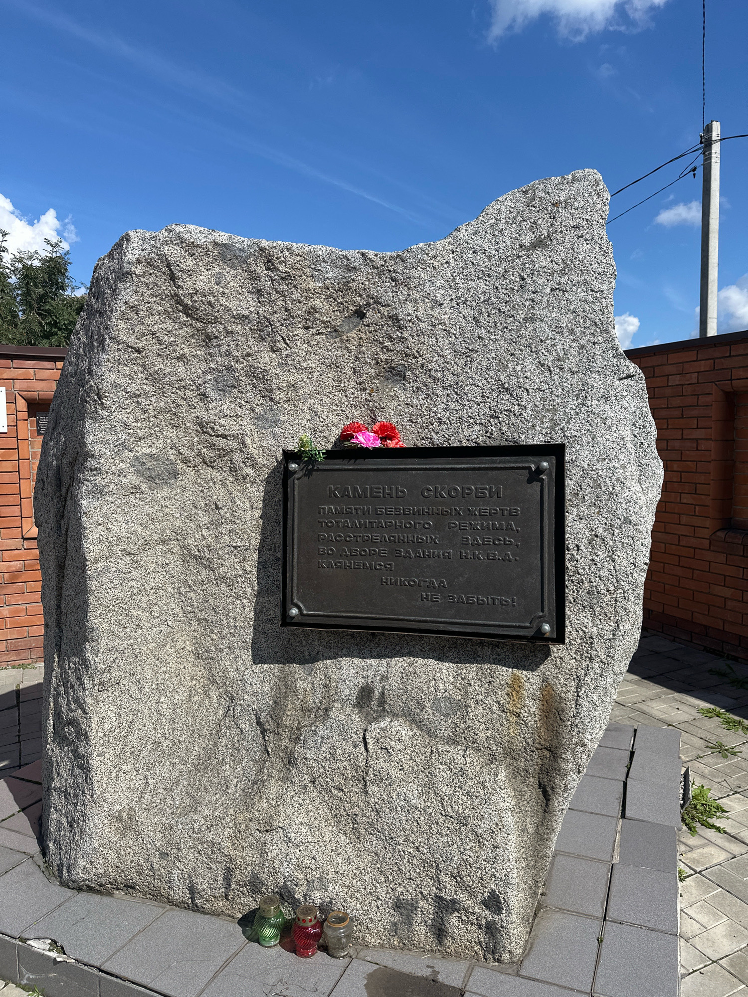 The image shows a stone monument with a dark metal plaque attached. The plaque has text inscribed on it and a bouquet of artificial flowers placed on top. Three small votive candles are arranged at the base of the stone. The monument is situated outdoors, with a brick wall and a clear blue sky with some clouds visible in the background. The ground around the monument is paved with grey tiles. The inscription on the plaque is in Russian and appears to be a memorial. Based on context from similar images and information about this specific monument, it is located in the city of Biysk, Russia. The monument is the "Stone of Sorrow," a memorial to the victims of political repression.