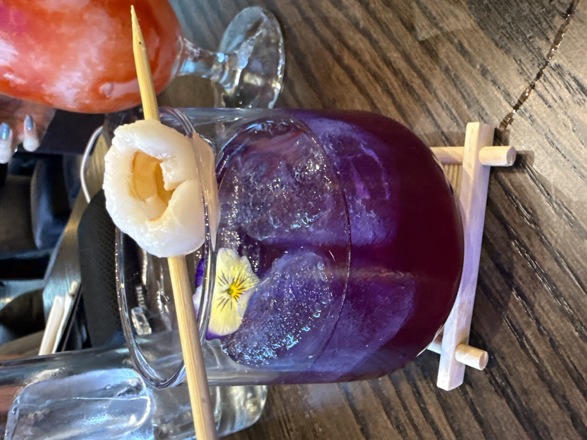 A still life of two cocktails on a wooden table. One cocktail is orange in color and the other is a deep purple, with ice cubes and a yellow flower visible inside the glass. A lychee fruit is skewered on a wooden stick and rests on the glass rim of the purple drink. A small wooden stand supports the side of the purple cocktail. A small part of a hand with silver painted nails is visible near the orange cocktail. Cutlery and other glassware are visible in the background. The location is San Bernardino Tlaxcalancingo, Mexico.