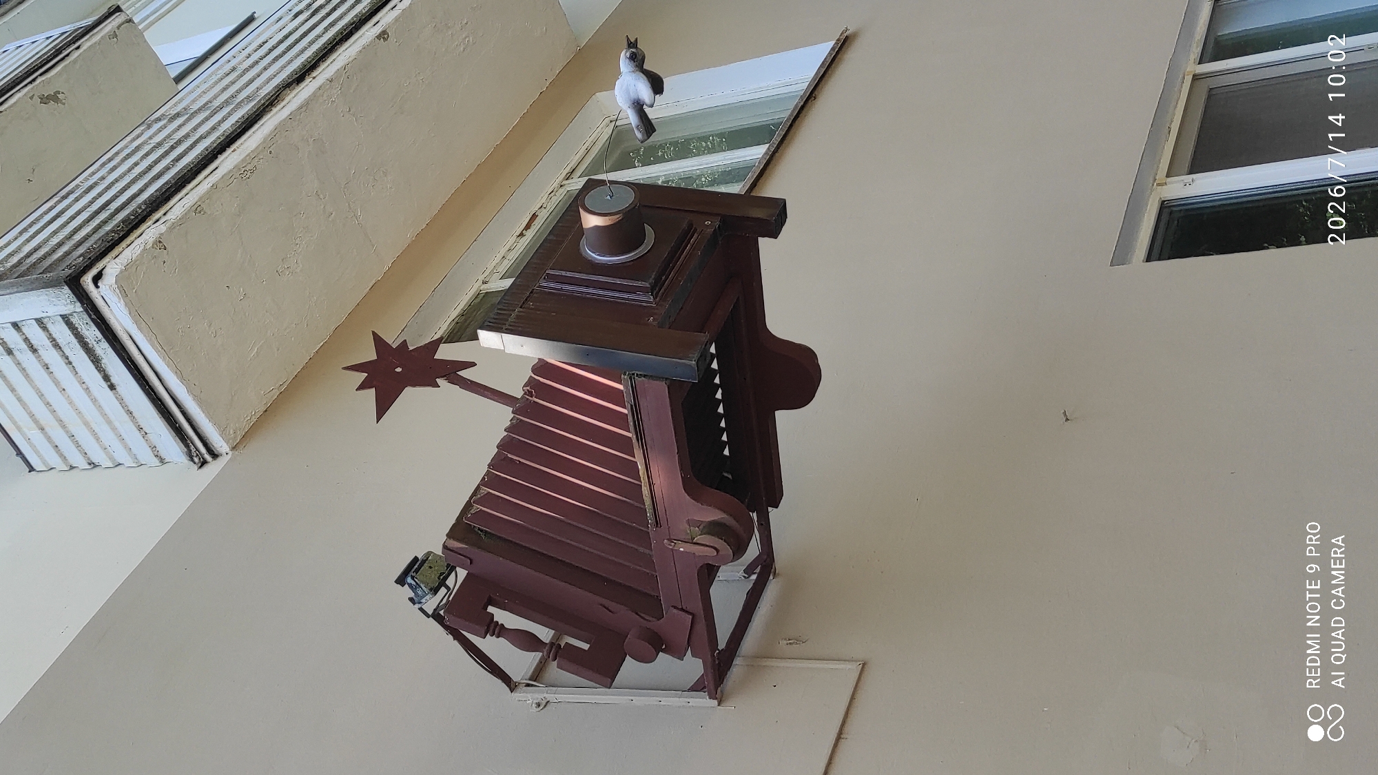 A large, stylized, red and brown antique camera sculpture is mounted outside a building window in Polatsk, Belarus. The sculpture, resembling a bellows camera, is attached to the building's exterior wall. A small, white, bird-like figure, possibly a sculpture or a prop, is suspended by a thin wire to the right of the camera, appearing to be in flight towards the camera's lens. The building has a cream-colored facade, and above the camera, a section of a balcony with white horizontal slats is visible, showing signs of age and weathering. A window with a light-colored frame is behind the sculpture, and trees are visible through the windowpane. The sky appears bright and sunny, suggesting daytime. The bottom right corner of the image displays a timestamp: "2026/7/14 10:02". Additionally, a watermark in the bottom left corner reads "REDMI NOTE 9 PRO AI QUAD CAMERA".