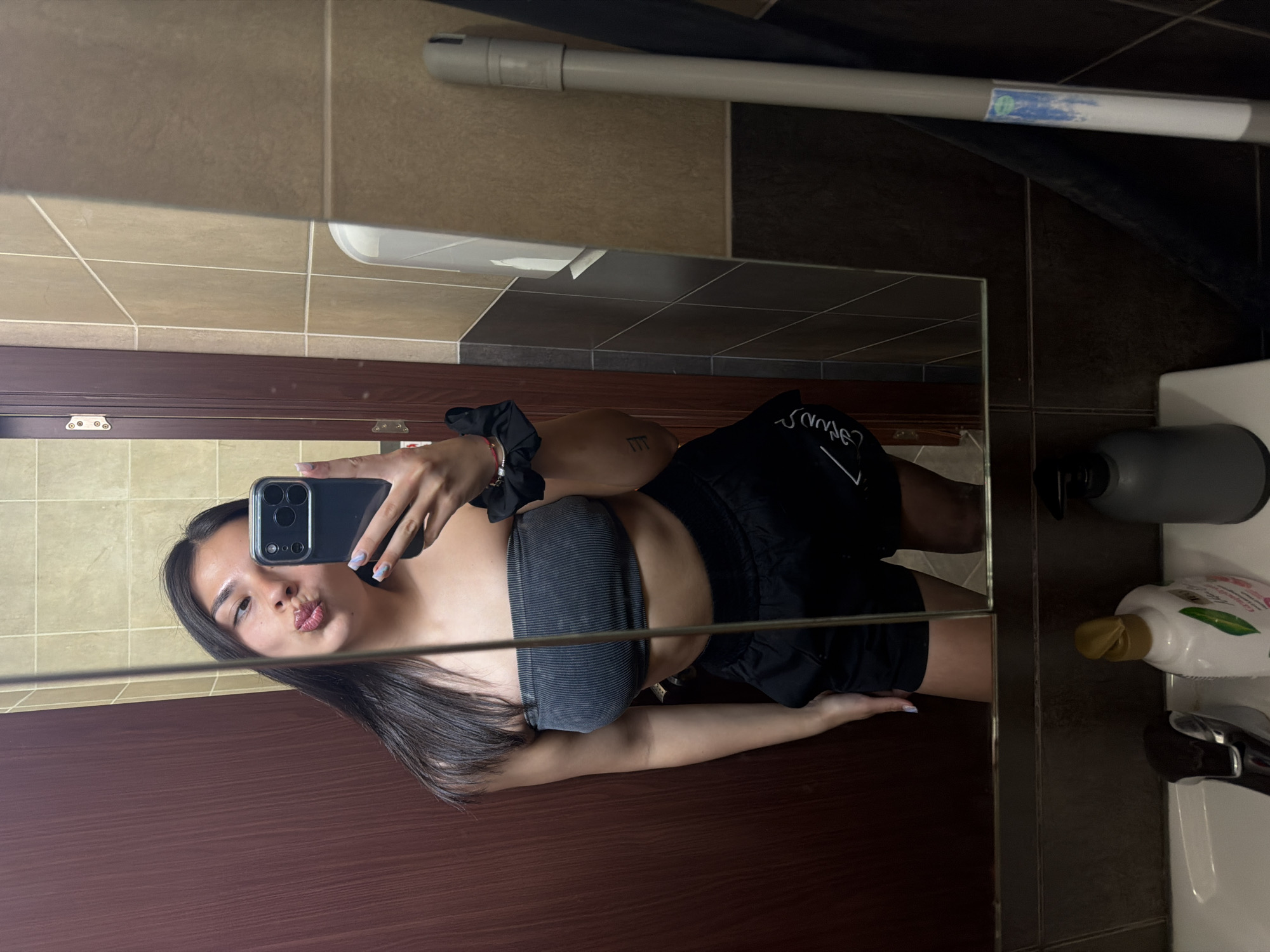 This rotated mirror selfie captures a young woman in a bathroom setting. The subject, with long dark hair and light skin, is reflected in the mirror, pouting her lips towards the camera. She is holding a black smartphone with a clear case in her right hand (as seen in the reflection) and her nails are painted a light color. She wears a dark grey or black ribbed bandeau top and dark shorts, which have some white, partially illegible text on them, possibly "SAVAGE". A black scrunchie is on her left wrist, and a thin red string or bracelet is visible on her right wrist. A small tattoo, resembling stylized double vertical lines, is visible on her right side below her armpit.
The bathroom features tiled walls with an upper section of light beige-brown square tiles and a lower section of darker brown/black square tiles, separated by a horizontal grout line. A dark wooden door or cabinet with visible hinges is reflected in the lower part of the mirror. On a counter to the right, a dark grey/black soap dispenser, a white sink basin, and a white bottle with a green leaf logo and gold cap are present. A light grey pipe runs across the upper part of the wall, bearing a partially faded blue and white label. The scene appears to be indoors with artificial lighting.