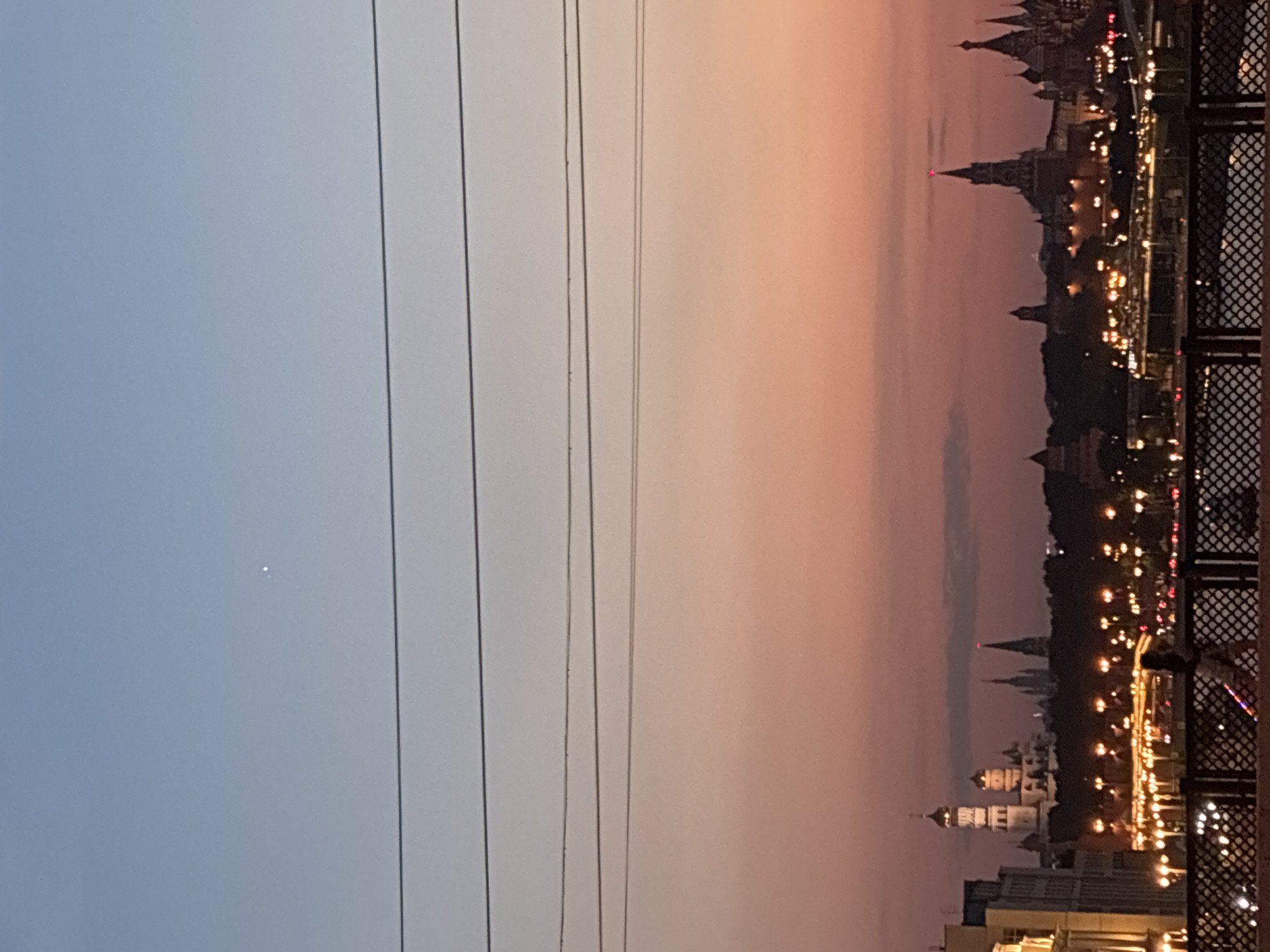 The image captures a cityscape at dusk or dawn in Moscow, Russia. Dominating the right side of the frame is the Kremlin's silhouette, illuminated with warm light. The buildings are partially obscured by trees, with a decorative metal railing in the foreground. Power lines extend across the left side of the image, where the sky fades from blue to peach. A solitary bright point is visible in the blue sky.
