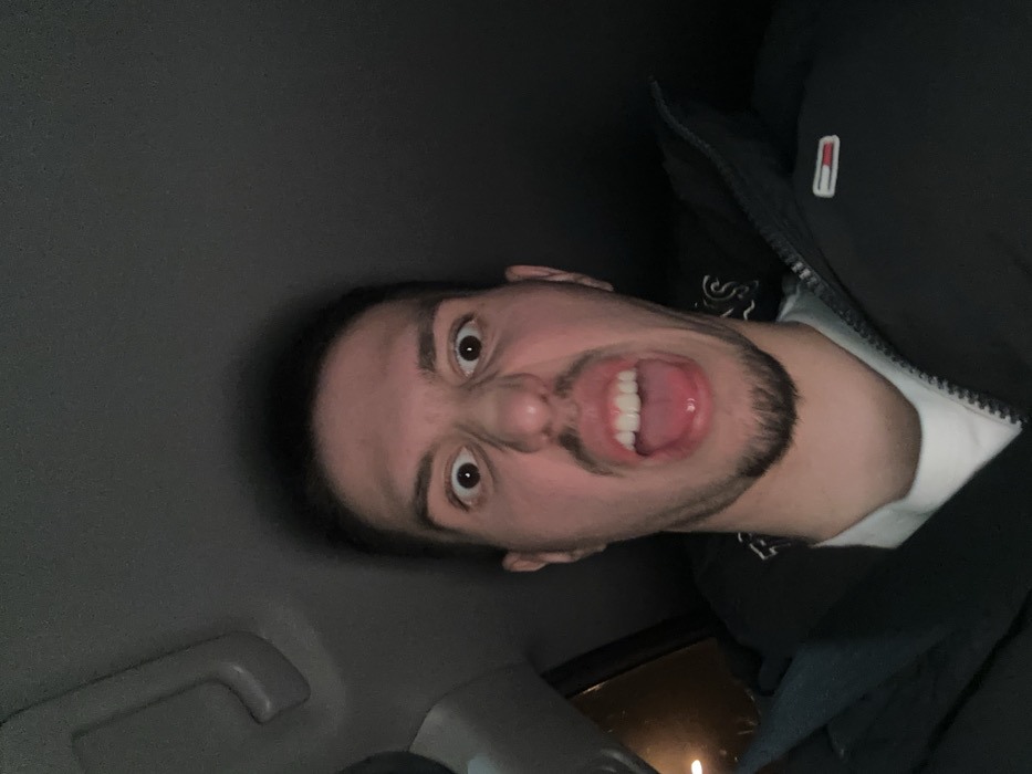 The image displays a male individual in a low-angle close-up, seated within the interior of a vehicle in Varna, Bulgaria. The subject possesses short dark hair, dark eyes, and a goatee. He is captured mid-expression, with his eyes wide open, eyebrows raised, nose wrinkled, and mouth agape, revealing both upper and lower teeth and a partially extended tongue. He is dressed in a dark, possibly padded jacket, worn over a white inner garment. A small rectangular badge, featuring horizontal red, white, and blue stripes, is affixed to the left chest area of his jacket. Overhead, the vehicle's dark gray headliner and a grab handle are visible in the upper left portion of the frame. The scene is dimly lit, indicative of a nighttime setting, with an indistinct warm light source visible outside the vehicle through a window on the left side of the image.