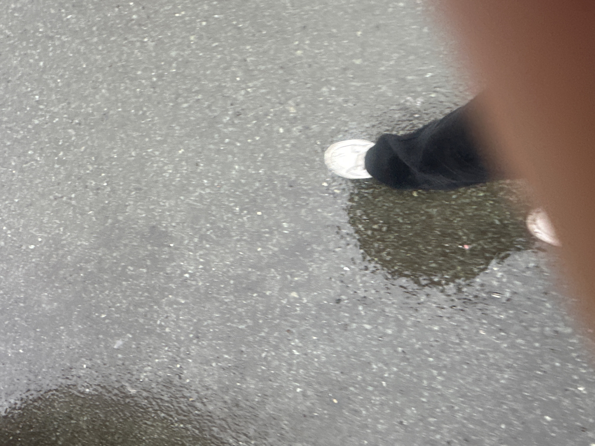 The image depicts a close-up, overhead view of a person's left foot wearing a white sneaker and black pants, stepping into a shallow puddle on a wet, asphalt surface. The asphalt is dark gray with visible small, light-colored aggregate particles. The puddle reflects the overcast sky and the surrounding environment. The lighting is dim, suggesting an outdoor scene under cloudy or rainy conditions. There is no identifiable urban context to place this in Petropavlovsk-Kamchatsky, Russia.