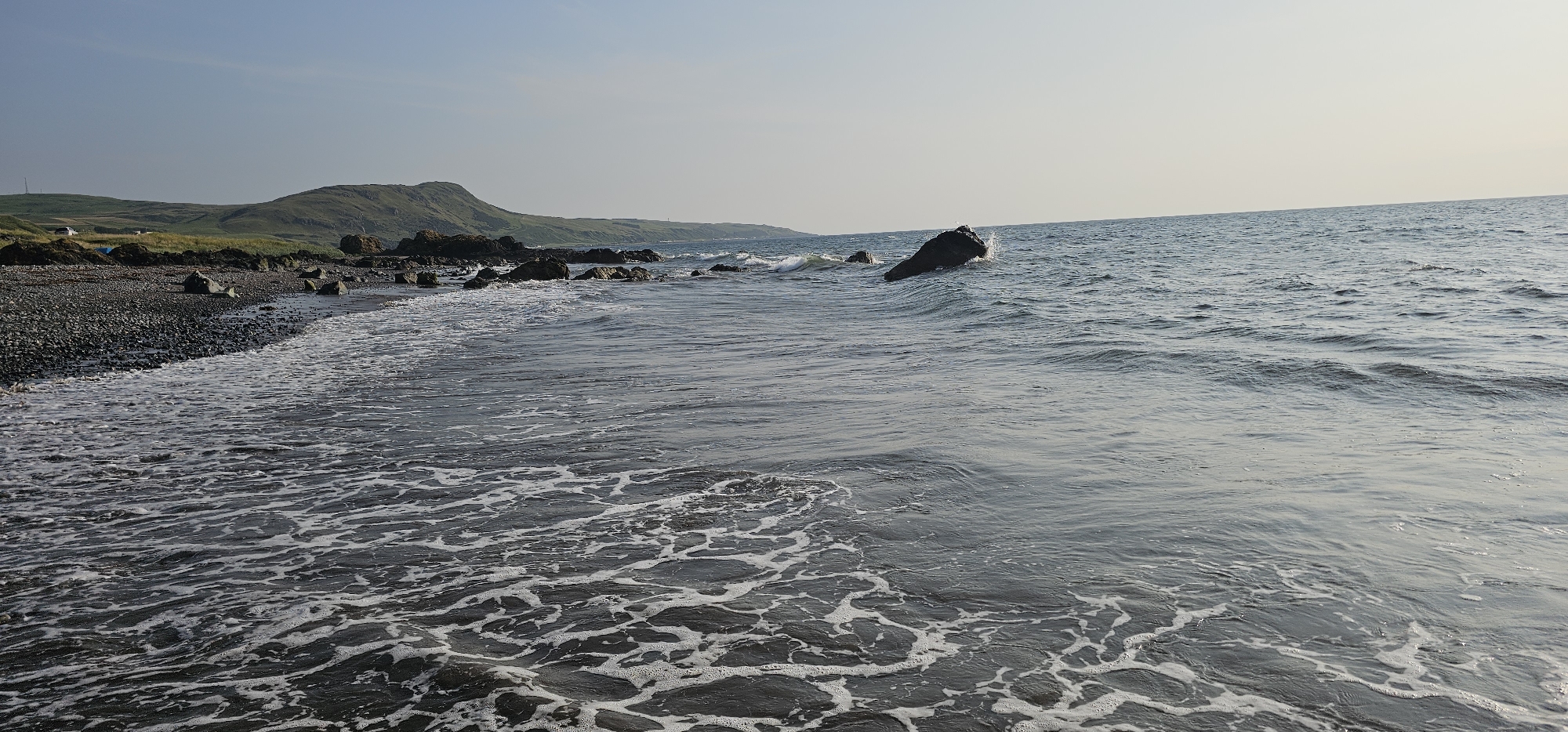 The image depicts a rocky beach with waves gently lapping at the shore. In the foreground, the water is foamy and turbulent, with a pattern of white froth on the dark surface. The beach itself is composed of dark pebbles and larger rocks. In the middle ground, larger rocks emerge from the water, and a prominent dark boulder stands against the incoming waves, with a splash of water visible. The background features rolling green hills that rise behind the coastline. The sky is a pale, clear blue, suggesting it is daytime. The scene is located on the coast of Girvan, United Kingdom. There are no people or animals visible in the image.