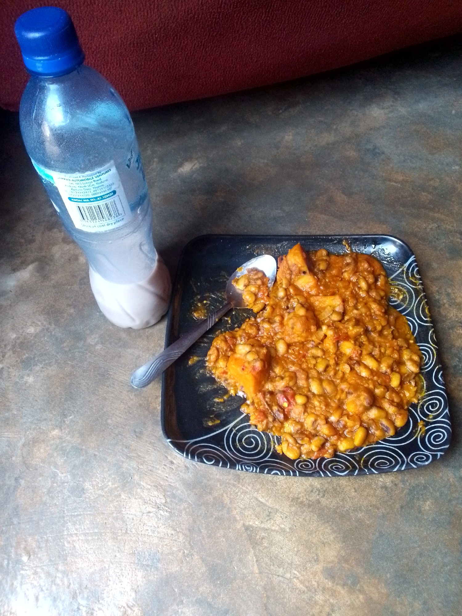 The image shows a plastic bottle of water next to a plate of food. The plate is black with white swirl patterns, and it contains a stew of beans and diced orange vegetables, possibly sweet potatoes. A metal spoon rests on the plate, partially submerged in the food. The bottle of water has a blue cap and a label with visible text, including "AUTOMOBILE VENTURES" and "Kaduna State Nigeria". The surface beneath the items appears to be a gray and brown textured floor. Part of a red-brown upholstered object is visible in the upper left corner of the image.