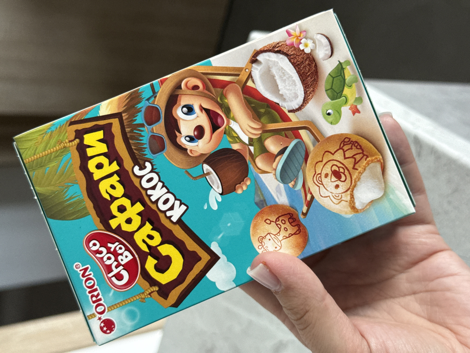 The image shows a close-up of a person's hand holding a box of Orion Choco Boy Safari Kokoc cookies. The box features a cartoon illustration of a monkey sitting in a beach chair, holding a coconut drink, with a halved coconut and tropical flowers nearby. The cookies themselves are round and decorated with animal images, including a giraffe and a lion. The background of the box is a bright blue sky with palm trees. The hand holding the box has light skin and well-maintained fingernails. The overall scene is bright and colorful, suggesting a product aimed at children or those seeking a tropical-themed snack. There is no discernible information in the image that would allow for the determination of its location within Lyubertsy, Russia.