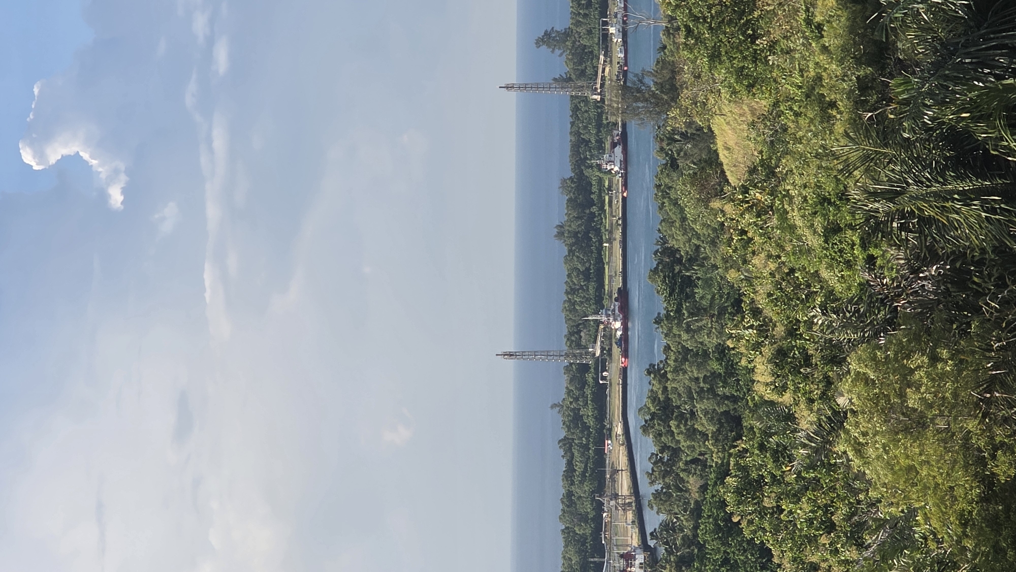 The image shows a channel of water bordered by land with dense green vegetation on the right and some structures visible along the land. Two tall, lattice-like structures are visible along the water. The sky is partly cloudy. The location is Lhokseumawe, Indonesia.