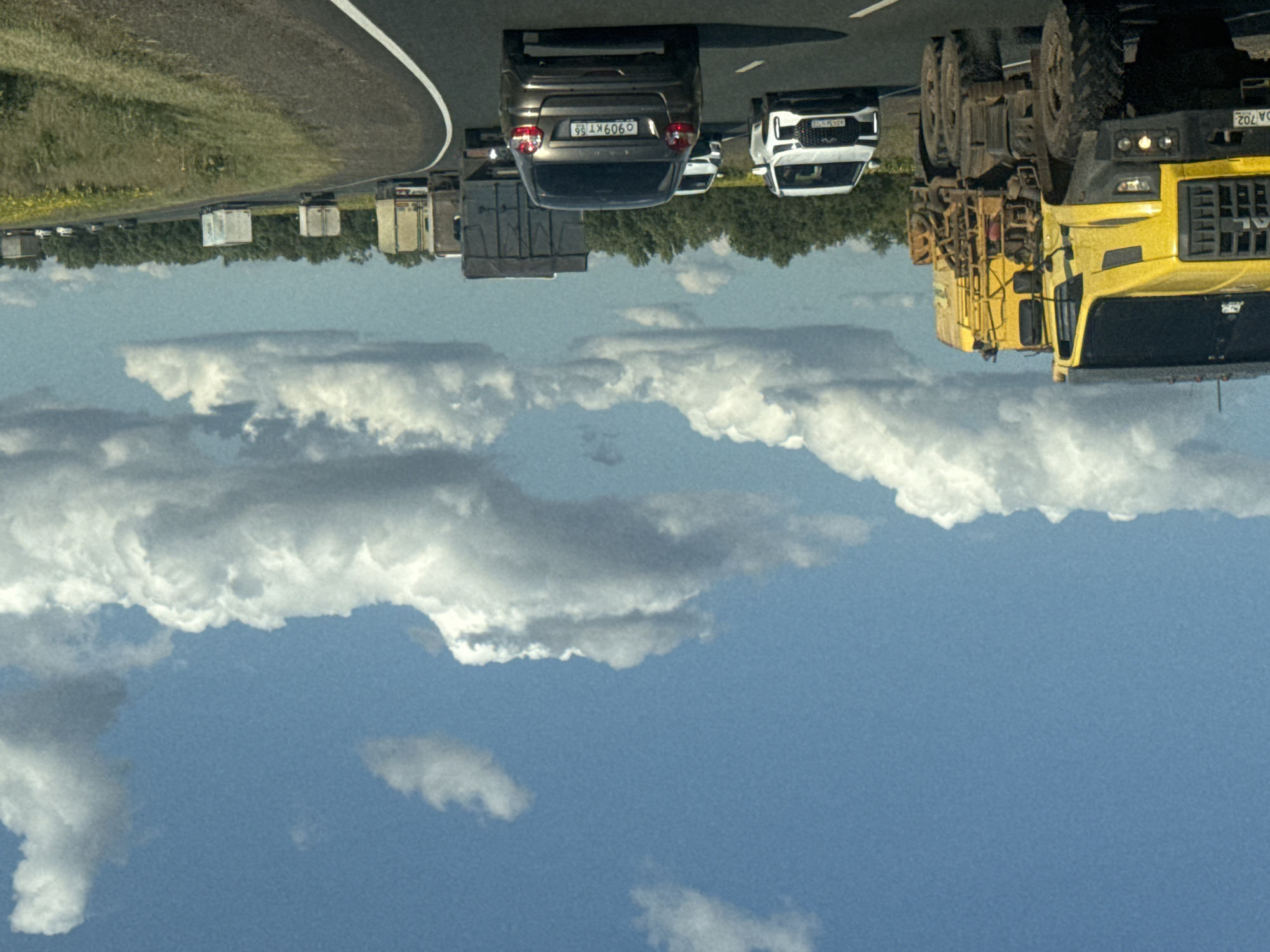 The image depicts a highway scene in or near Perevolotsky, Russia, under a bright blue sky with scattered white clouds. A large yellow truck, possibly a specialized construction or utility vehicle due to its bulk and attached equipment, is in the foreground on the left. It has large, rugged tires and a license plate visible that appears to read "DA 702". Several other vehicles are on the road, including a white SUV on the right of the yellow truck, followed by a dark sedan with the license plate "O 909 KT 56". Further down the highway, a line of trucks with various trailer types, including box trailers and flatbeds, are visible traveling in the same direction. The road is paved, with white lane markings. The surrounding landscape features a dense line of green trees on the right, and a field with yellow flowers, likely sunflowers, bordering the road. The overall impression is a sunny day on a rural or semi-rural highway in Russia.
