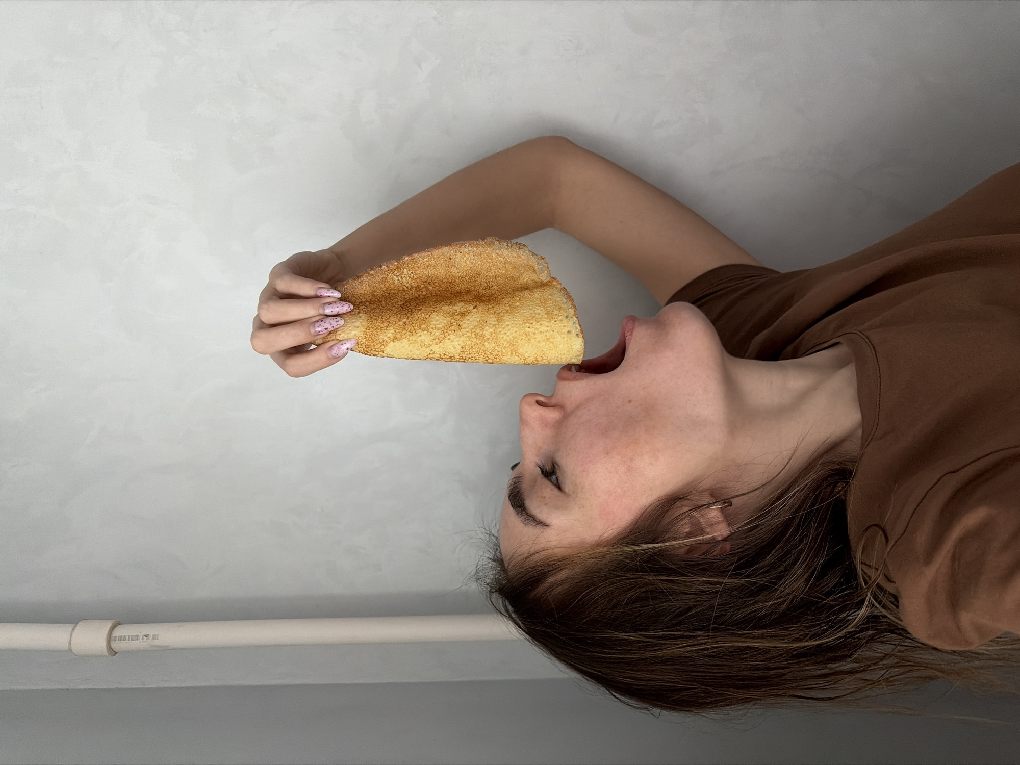 A woman is holding a folded, cooked pancake towards her open mouth. She is lying on her side with her head tilted back, and the pancake is positioned as if she is about to take a bite. The woman has fair skin, light brown hair with blonde streaks, and painted fingernails. She is wearing a brown t-shirt and gold hoop earrings.
The background is a light grey wall with a white pipe running along the lower section. The image is rotated. Based on the information provided, it is not possible to determine if the photo was taken in the city of Neftekamsk, Russia.