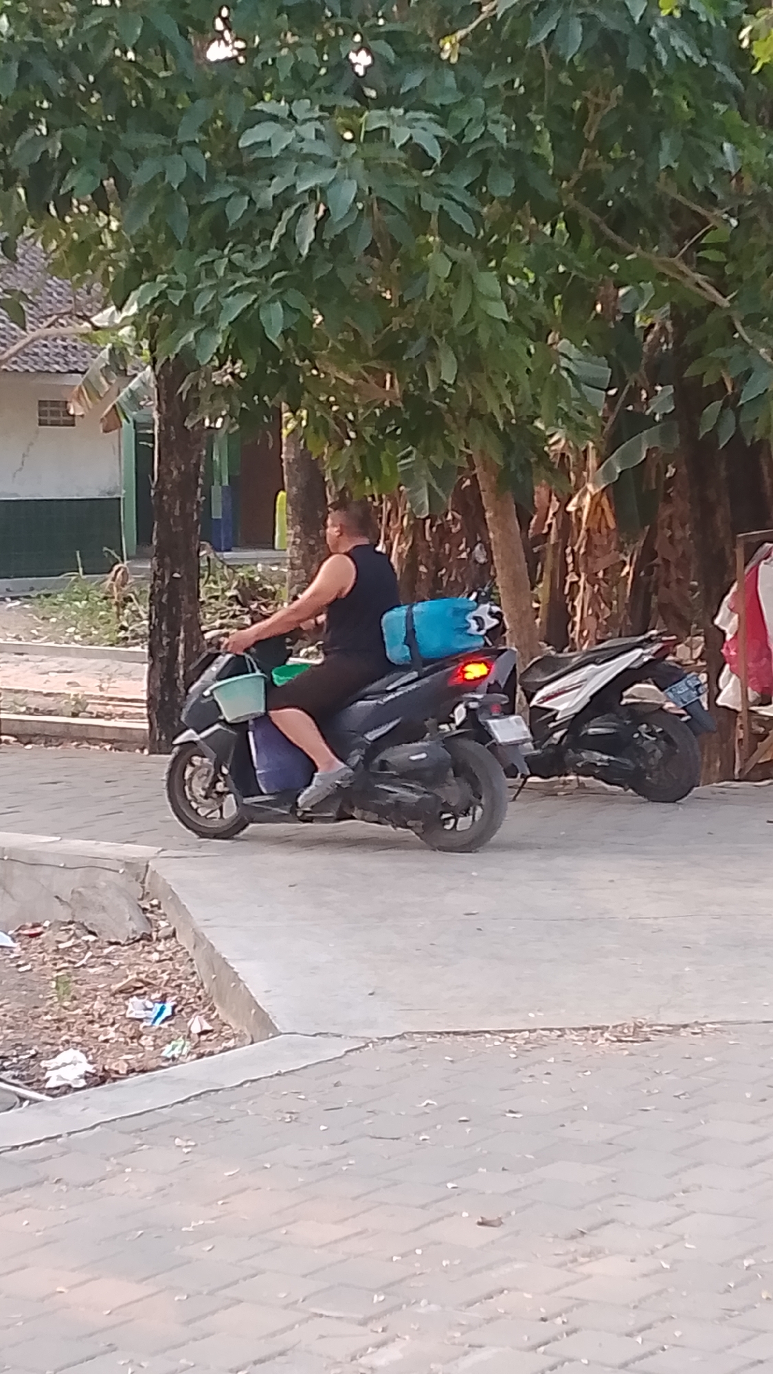A person is riding a dark-colored scooter down a paved road. They are wearing a black tank top and black shorts, with gray shoes. The scooter has a light blue duffel bag strapped to the back and a light green bucket on the front. There is another scooter parked behind it, which is white and black. To the left of the road is a ditch filled with debris, and beyond that, a building with a tiled roof and a green wall. Tall trees with green leaves line the right side of the road, and a banana tree is visible in the background. The ground is paved with light gray bricks. The scene appears to be outdoors during daylight.