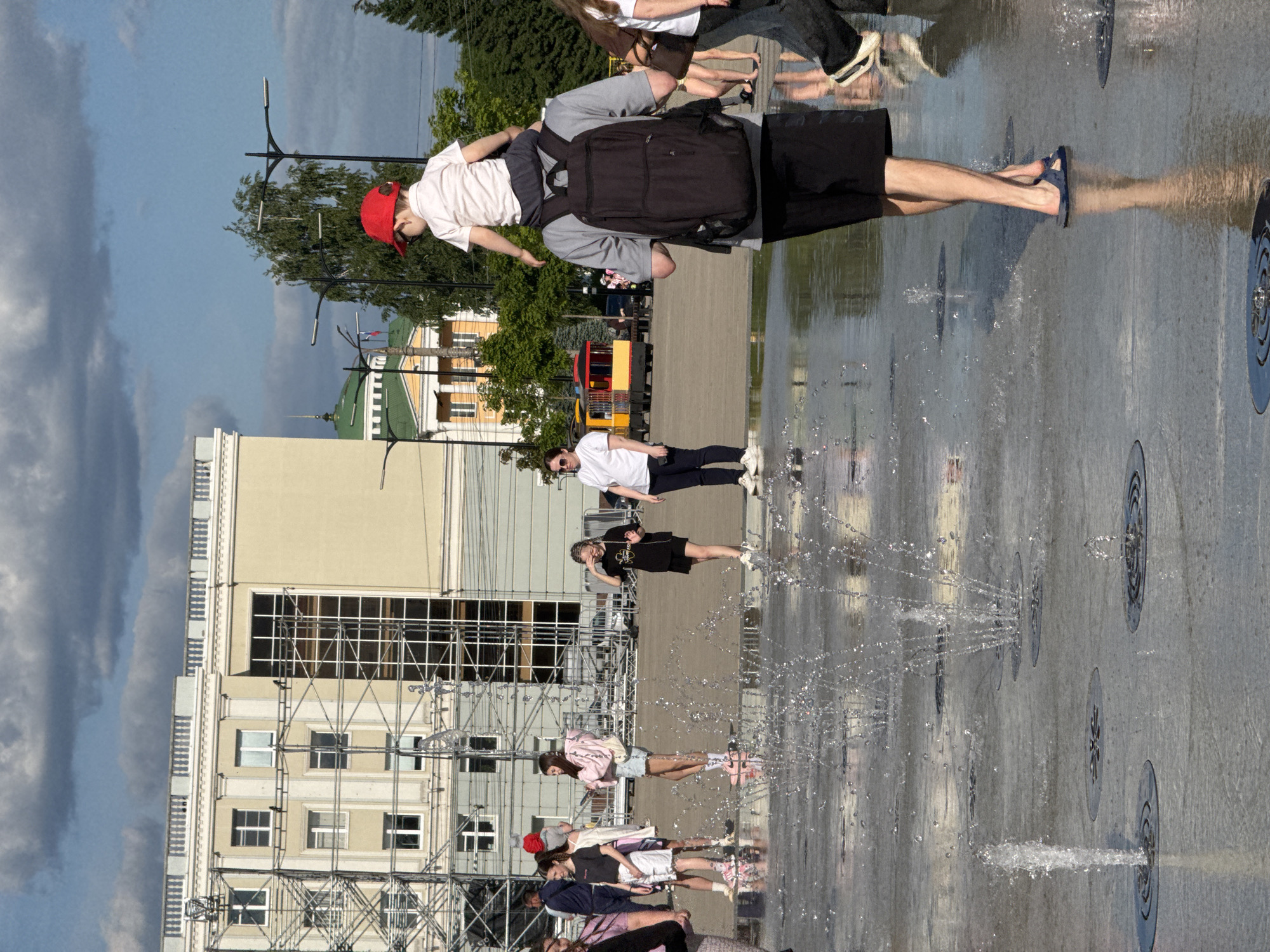 The image captures a sunny day in a public square in Izhevsk, Russia. In the foreground, a ground-level fountain with multiple water jets erupts from circular grates embedded in a wet, light-colored paved surface. A man, viewed from the back, walks through the fountain area wearing a grey t-shirt, black shorts, and blue flip-flops. He carries a black backpack and a small child on his shoulders, who wears a white shirt and a red baseball cap.
In the mid-ground, several individuals are present around the fountain. A young woman in a pink hoodie and denim shorts stands to the left, partially turned away. Another young woman in a black t-shirt and black shorts shields her eyes with one hand. Further left, a woman in a white t-shirt and dark pants stands facing right.
The background features a large, light beige building with white trim and a balustrade along its roofline. Extensive metal scaffolding is erected against its left side, partially covering a prominent glass section. Further back and to the right, a building with a green dome, yellow walls, and white columns is visible. Multiple black streetlights and green trees are interspersed throughout the scene. A yellow and red open-sided tourist train is parked or moving slowly in the mid-background. The sky is partly cloudy with areas of blue visible.