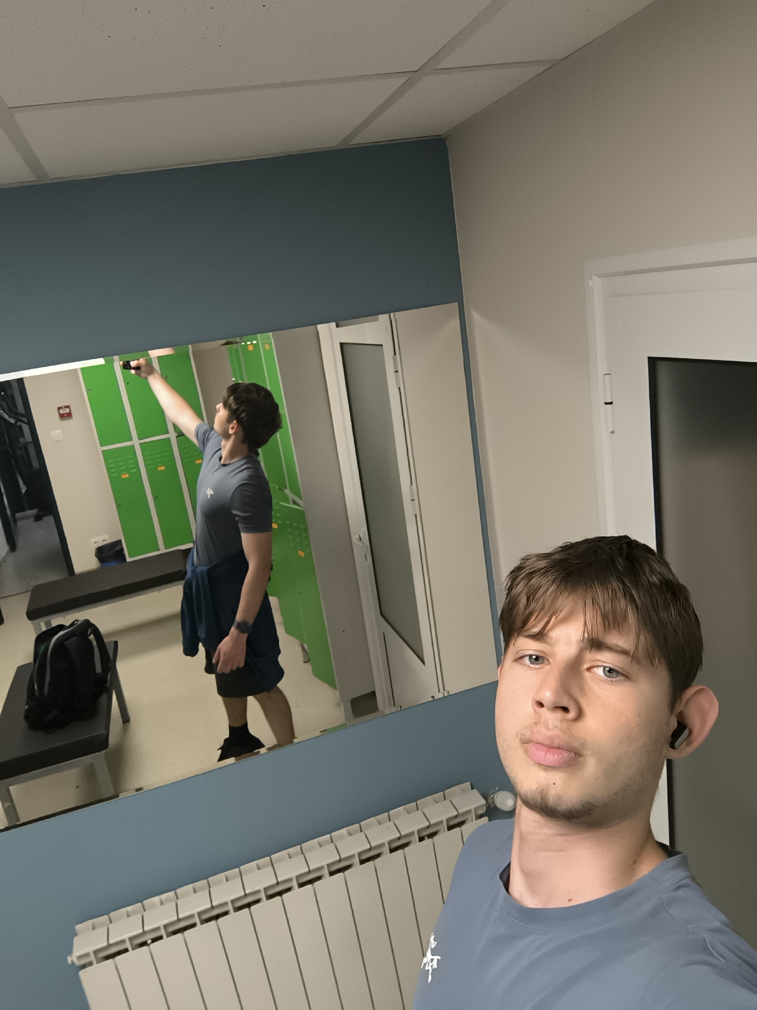 A young man takes a selfie in what appears to be a locker room in Sofia, Bulgaria, indicated by the Cyrillic writing on a red emergency information placard reflected in the mirror. The room contains green lockers, a bench with a black backpack on it, and a radiator below the mirror. The man in the foreground wears a gray t-shirt, an earbud in his right ear, and is positioned near a white door. The mirror reflects the man taking the photo and the locker room behind him.