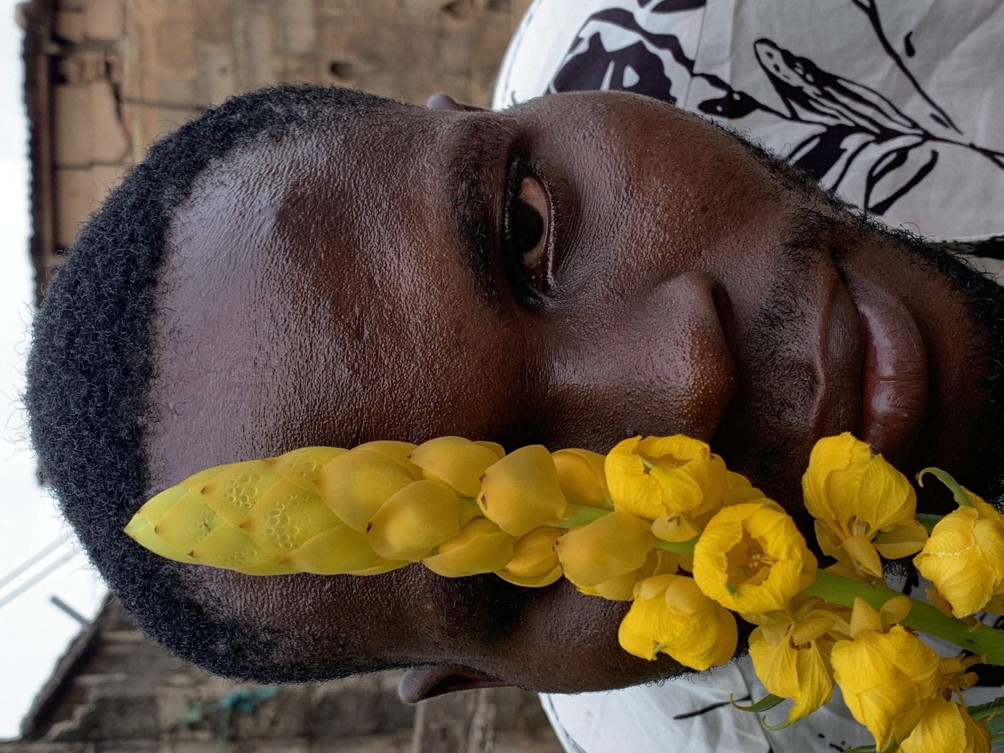 A close-up portrait shows a dark-skinned man with tightly coiled black hair, holding a stem of yellow flowers against his face. The flowers appear to be candlestick senna. The background is blurred and shows a wall and a shirt with black and white patterns, along with a stairway. The location might be in Ikorodu, Nigeria based on the prompt.