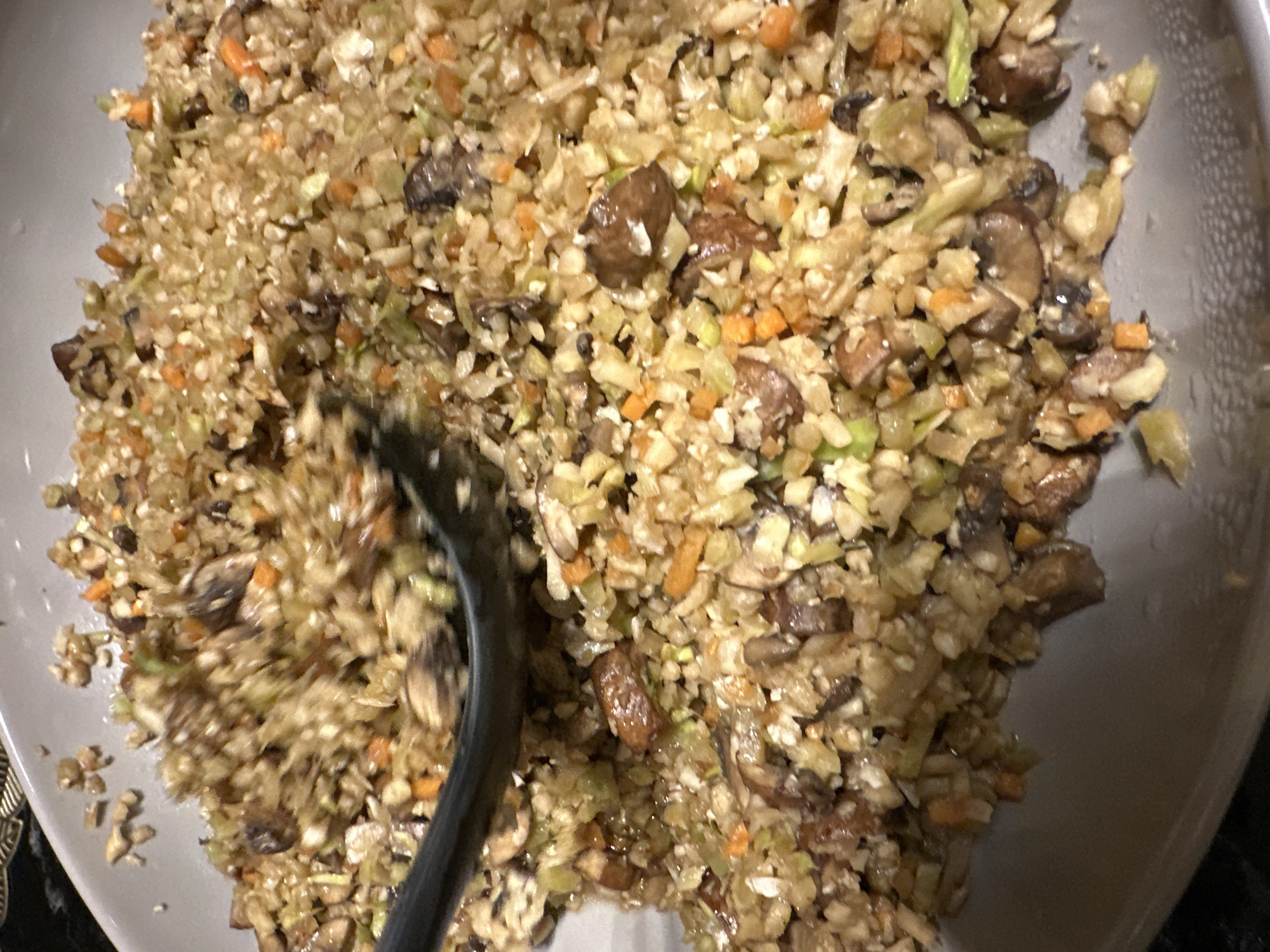 A close-up, high-angle shot shows a bowl of cooked food being stirred with a black utensil. The food appears to be a mixture of ingredients, including chopped mushrooms, shredded cabbage or cauliflower, and diced carrots, all lightly coated in sauce. The bowl is a light gray, and the scene is dimly lit. No information in the picture indicates Mexico City.