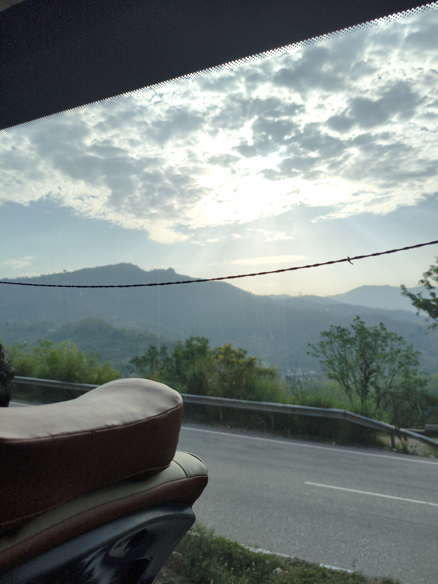 A medium shot captures a motorcycle seat in the foreground, viewed from the rear and side. The seat is brown with darker brown trim. Behind the motorcycle, a two-lane asphalt road curves to the right, bordered by a metal guardrail. Beyond the road, lush green hills and mountains recede into the distance, fading into a hazy blue. The sky above is filled with scattered white and grey clouds, with sunlight breaking through in rays, indicating it is likely early morning or late afternoon. A thin, dark wire stretches horizontally across the middle of the frame, obscuring part of the distant mountain range. The scene suggests a scenic route, possibly in the outskirts of Bilaspur, India, given the mountainous terrain and vegetation.