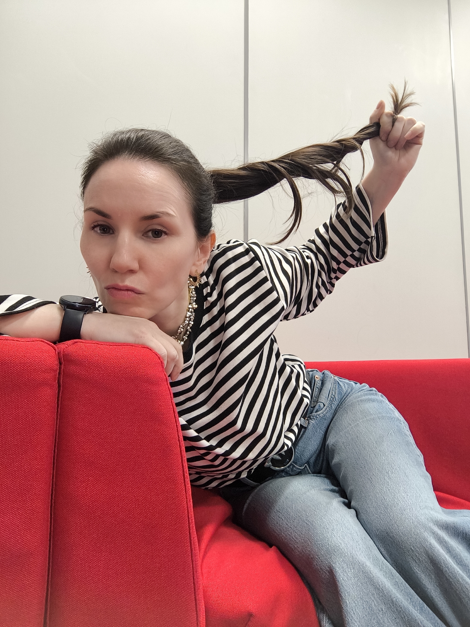 A woman with brown hair pulled back in a ponytail is lying on a red sofa. She is wearing a black and white striped long-sleeved shirt and blue jeans. Her left arm is resting on the sofa, with a black smartwatch on her wrist. Her right hand is holding the end of her ponytail. She is looking at the camera with a slight frown. Behind her is a white wall. Location: Unknown, Unknown.