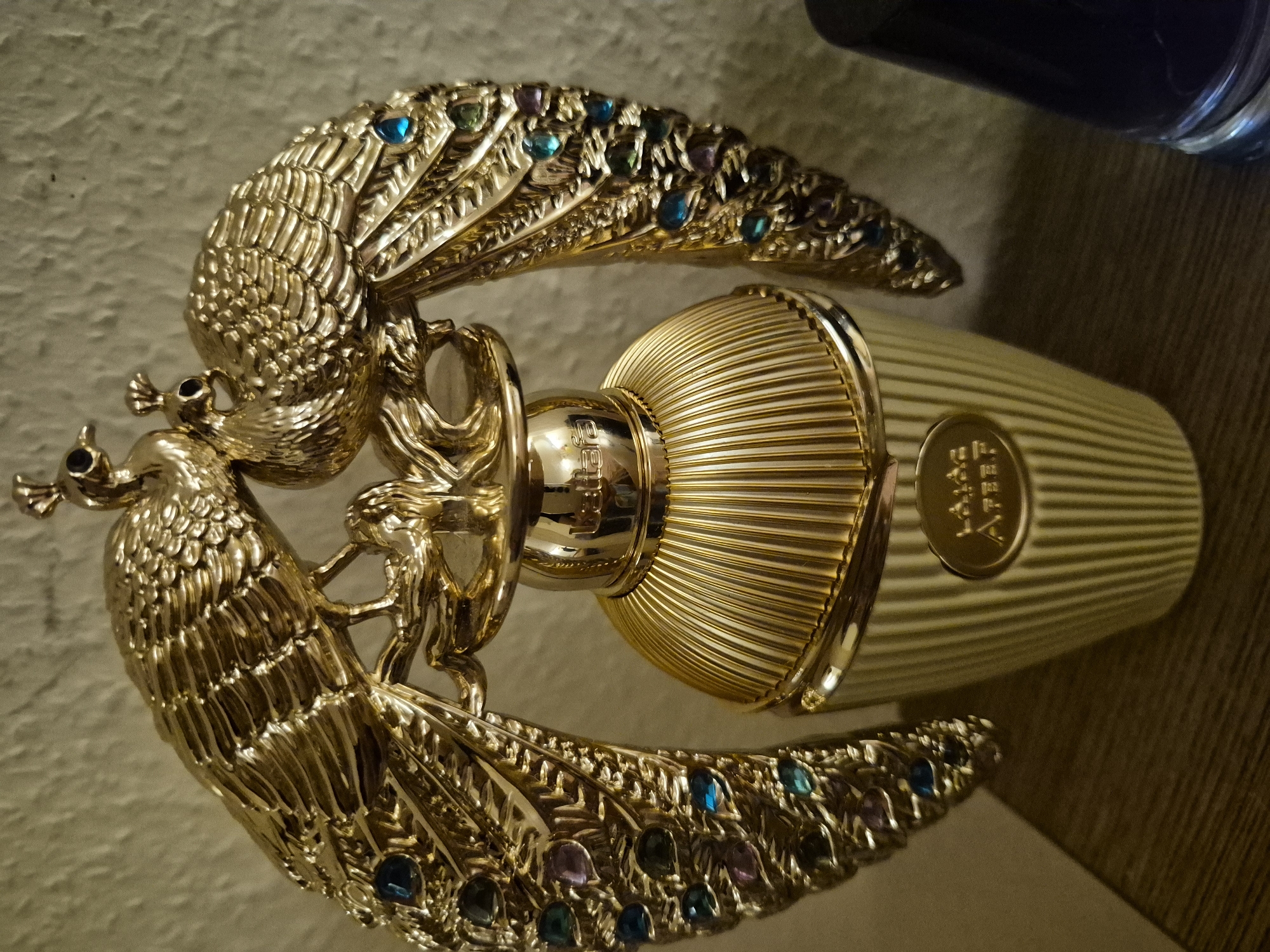 A close-up, eye-level, medium shot focuses on a golden perfume bottle with two ornate peacocks adorning the top. The bottle is ribbed and has a ribbed cap. The peacocks are intricately detailed, with their feathers fanned out and embellished with small, colorful gemstones in shades of blue, green, and purple. One peacock is positioned slightly behind and above the other. The background is a textured, light-colored wall, and the perfume bottle is sitting on a dark, wooden surface. The overall lighting is warm and highlights the metallic sheen of the bottle and peacocks.