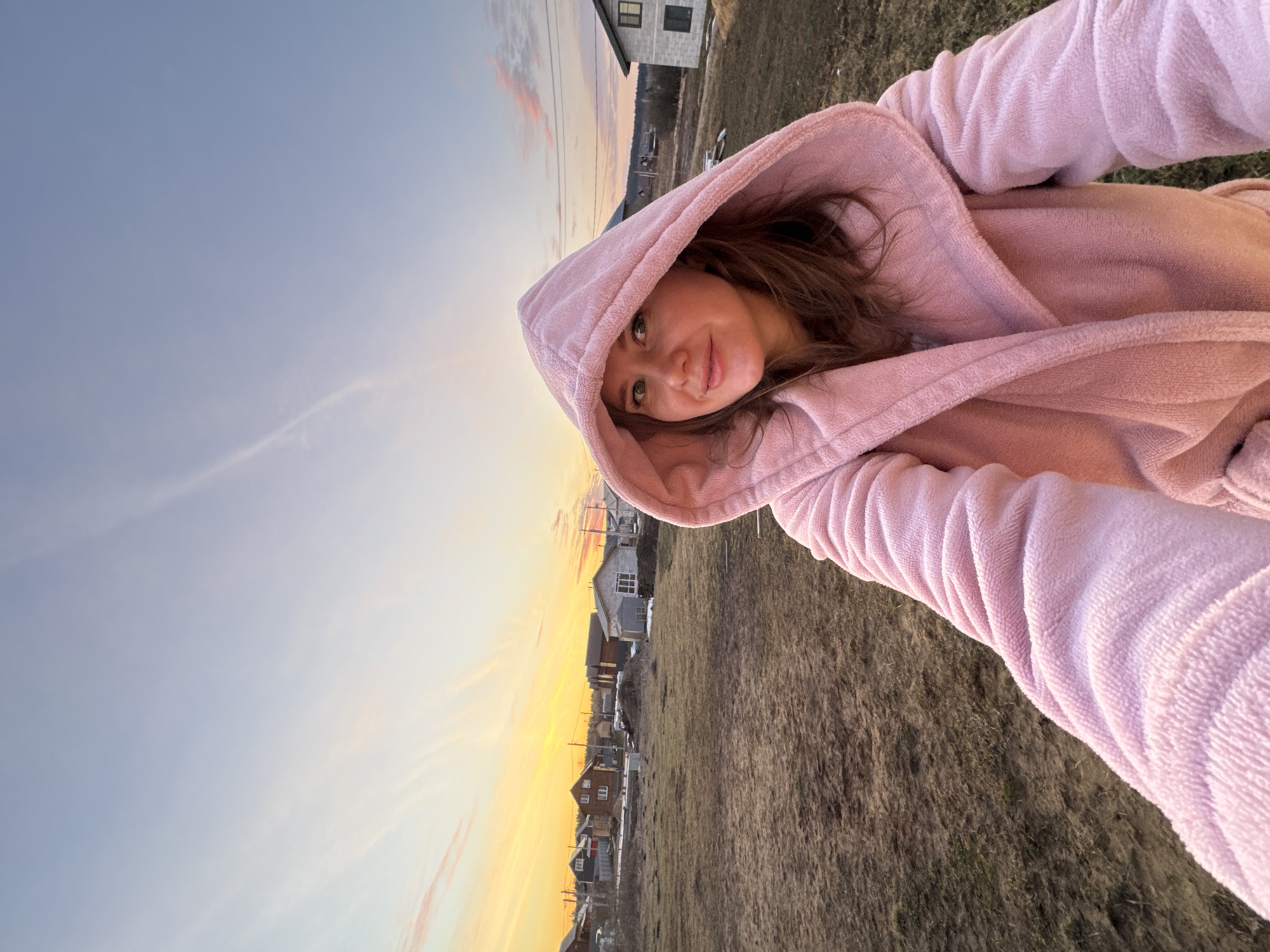 A woman wearing a pink hooded robe is taking a selfie outdoors at what appears to be dusk or dawn. The sky transitions from blue to orange in the background. In the distance, there are houses, and the foreground shows a field with dry grass. The location seems to be near a residential area, possibly in Gornyi (Dvurechenskoe S/P), Russia, based on the context.