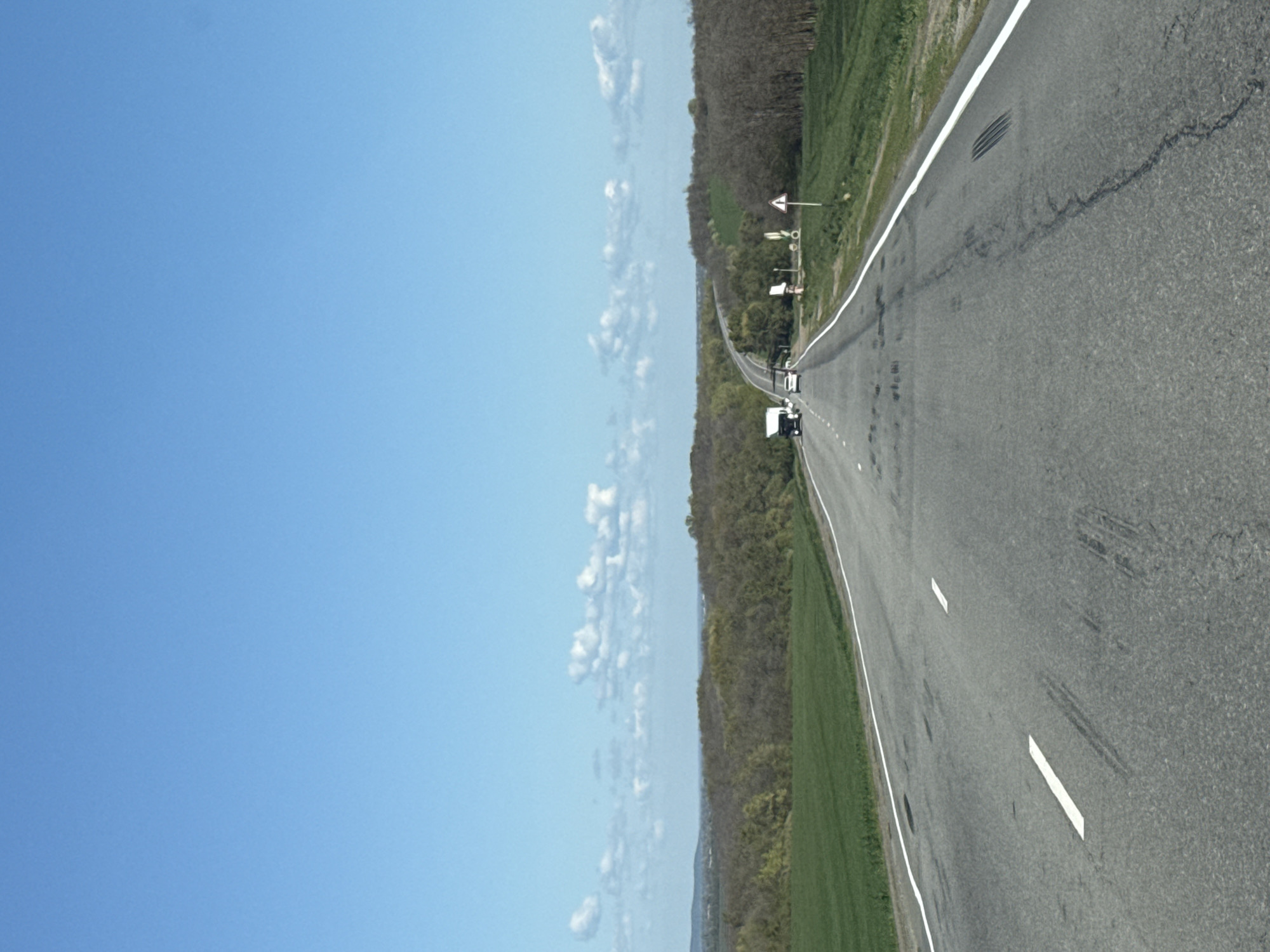 The image shows a straight asphalt road in Pokrovskoye, Russia, flanked by a landscape of trees and green grass on one side and a clear blue sky with scattered clouds on the other. A few vehicles are seen driving on the road in the distance. Road markings, including white lines, are visible on the road surface.