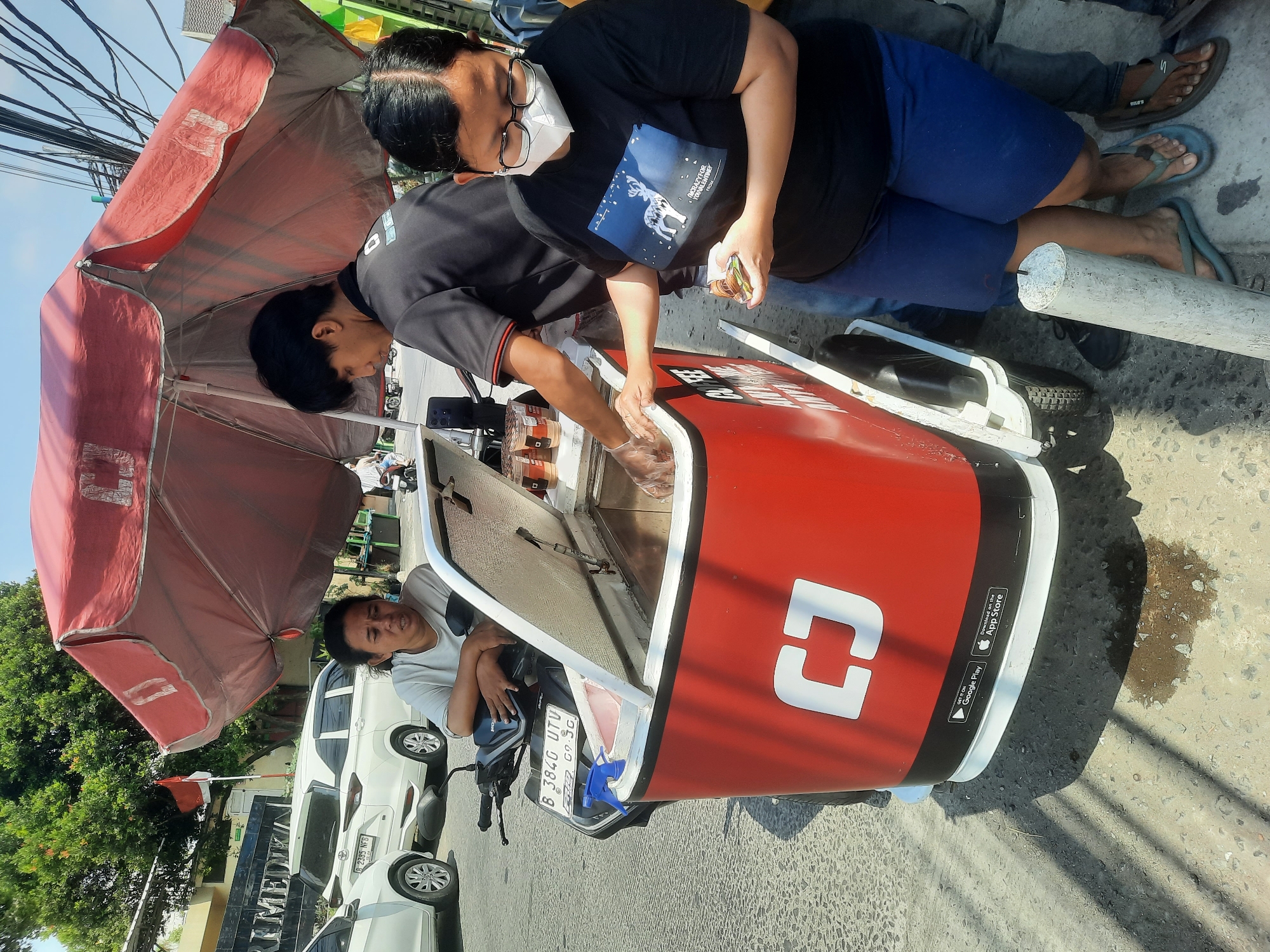 In North Jakarta, Indonesia, on a sunny day, a mobile beverage vendor operates from a red and white cooler attached to a motorcycle. The vendor, a man with black hair wearing a black polo shirt, is seen serving a customer. The customer, a woman wearing glasses and a white face mask over her nose and mouth, is dressed in a black t-shirt with a deer graphic and blue shorts. She is holding a small item in her hand. Another woman, wearing a white t-shirt, is seated on the motorcycle, appearing to be part of the operation. The surrounding environment shows a paved road, parked white cars, and electrical wires overhead. The red umbrella shielding the vendor and customer features a white logo. The side of the cooler has a white logo and app store download icons.