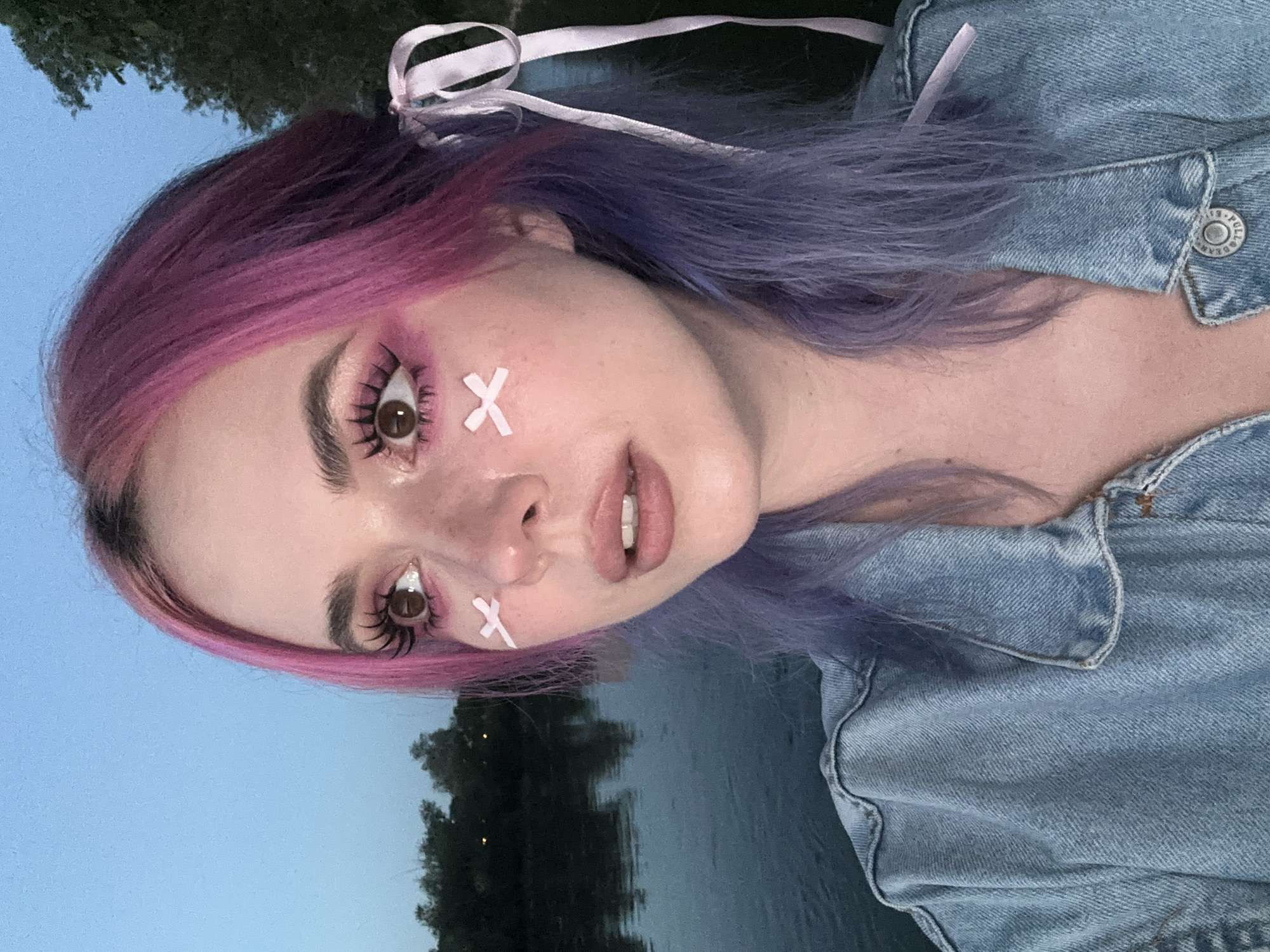 A close-up shot shows a person with pink and purple hair, wearing a denim jacket. Two white "X" marks are visible on their cheeks, near the outer corners of their eyes. Their eyes are enhanced with pink eye shadow and long lashes. They are wearing a pink ribbon in their hair. The background depicts a body of water and trees. The location cannot be determined without further information.