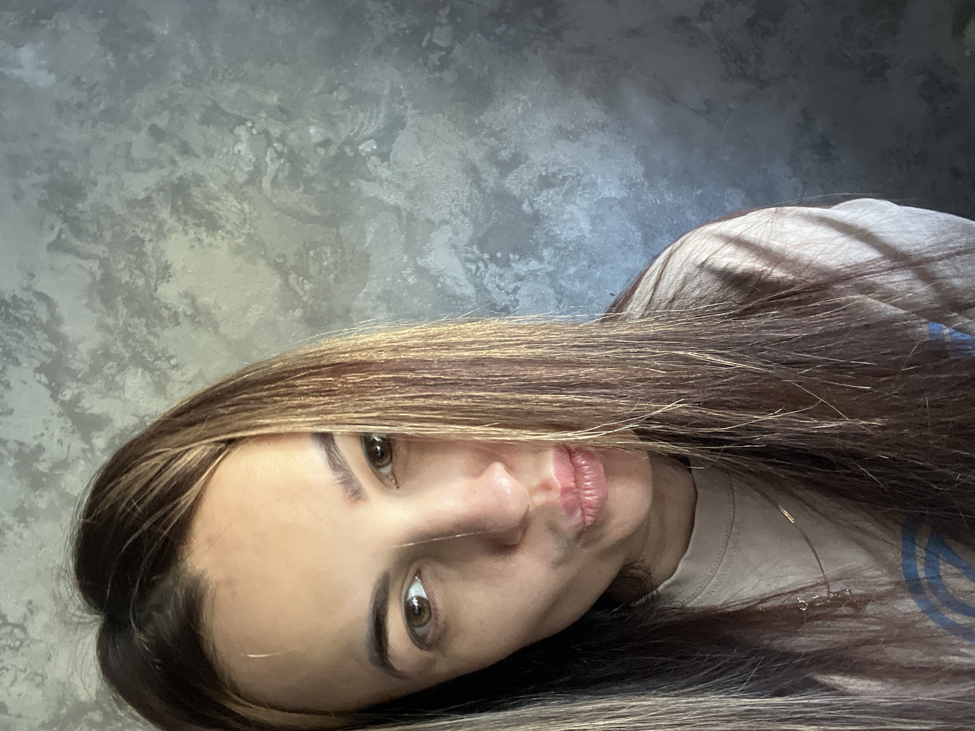 A selfie of a woman with long, brown hair with blonde highlights, posing against a gray mottled background. The woman's face is tilted to the side, with her eyes open and looking towards the camera. She has fair skin and lightly colored lips. A thin necklace is visible around her neck, and she appears to be wearing a light-colored t-shirt. The location and city are unknown.