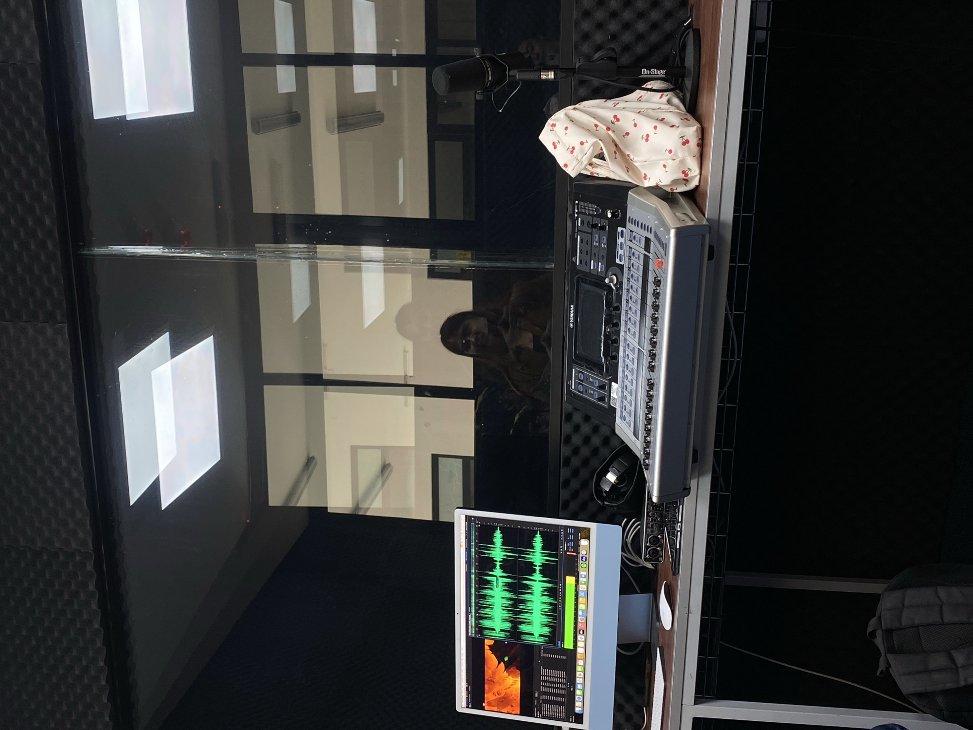 The image shows an indoor recording studio with soundproofing material on the walls. Visible equipment includes a computer monitor displaying waveforms, a mixing console (Yamaha), a microphone (Shure), and headphones. A patterned cloth (cherries) is draped over the console. The reflection in the glass reveals a person. The location may be in Puebla, Mexico.