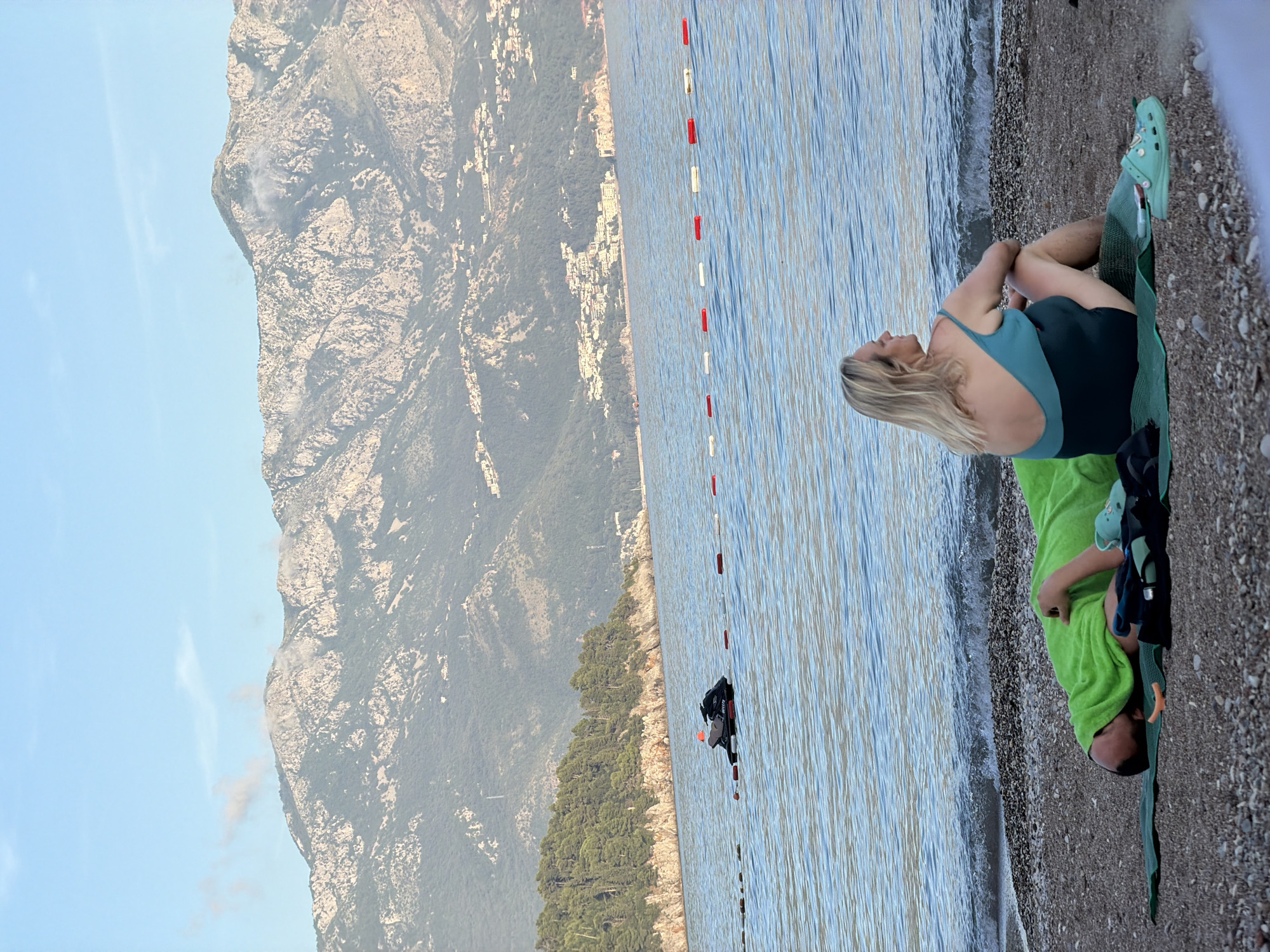 The image, correctly oriented, depicts a serene coastal scene, characteristic of Budva, Montenegro, with majestic mountains meeting the sea. In the foreground, a woman with blonde hair, wearing a teal and dark blue swimsuit, sits on a green mat on a pebble beach. She is positioned with her back partially to the viewer, looking contemplatively towards the tranquil water and mountains. Her arms are crossed, and she wears light blue Crocs. Beside her, a child lies on a bright green towel, partially obscured, also facing the water.
The sea is calm, with gentle ripples near the shore, suggesting pleasant weather and a peaceful atmosphere. A line of red and white buoys extends across the midground water, indicating a designated area. Further out, a person on a dark personal watercraft, possibly a jet ski, is visible in the water.
The background is dominated by dramatic, rocky mountains covered in sparse vegetation, rising steeply from the coastline. Small buildings and settlements are nestled on the lower slopes of these peaks. The sky is clear blue with a few faint clouds, and the soft lighting suggests either early morning or late afternoon. The overall impression is one of relaxation and natural beauty at a scenic beach destination. No visible text is discernible in the image.