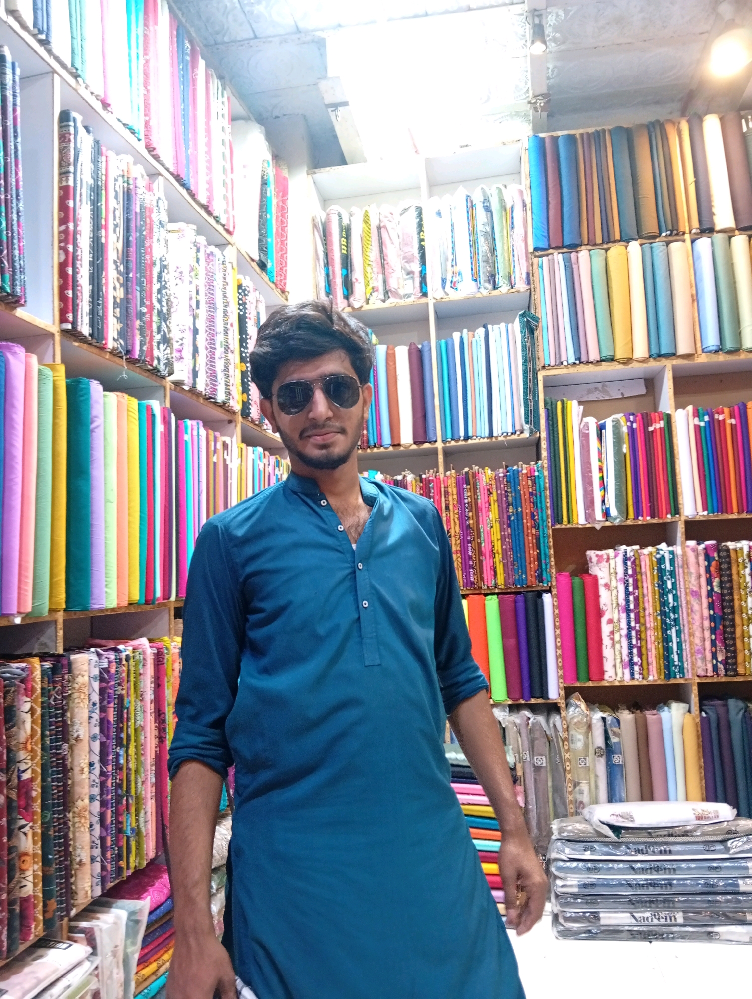 In Larkana, Pakistan, a man wearing a blue kurta and sunglasses stands in a fabric store. The store is filled with shelves stocked with rolls of colorful fabric, displaying a variety of patterns and colors. The man is positioned in front of a wide selection of fabrics, with more rolls visible on shelves behind him, extending into the background of the shop. The lighting in the store appears to be artificial, with overhead lights illuminating the scene.