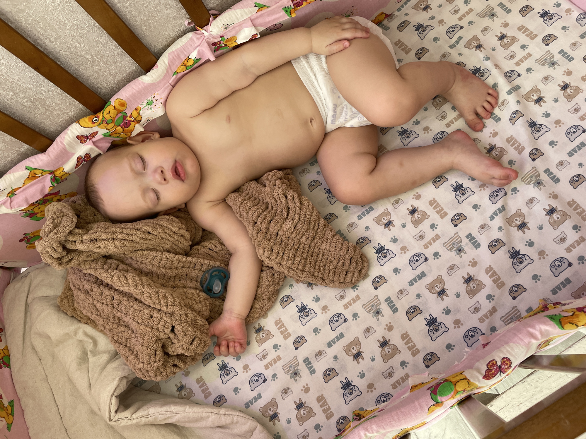 A baby is sleeping in a crib with a brown blanket. The baby is wearing a white diaper and has a pacifier next to their hand. The crib has a bear-themed pattern on the bedding and a padded bumper with cartoon animals on the sides. The location is Perm, Russia.