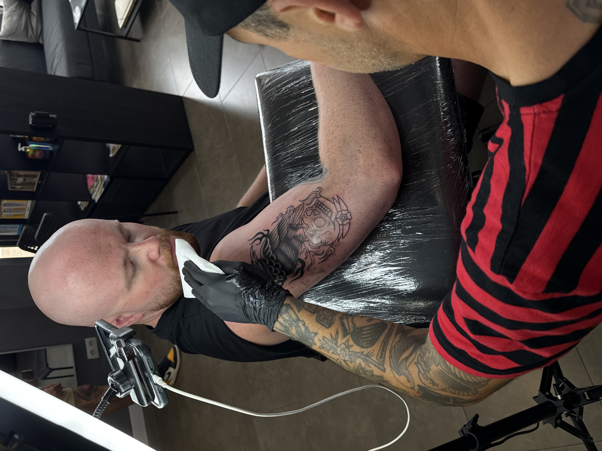 A tattoo artist in Yaiza, Spain, is in the process of applying a tattoo to a client's upper arm. The client is a bald man with a reddish beard, wearing a black t-shirt, and has a mobile phone propped up on a stand next to him. The tattoo is a large black and white design with elements of a skull. The tattoo artist, wearing a black baseball cap and a red and black striped shirt, has heavily tattooed arms and is focused on the client's arm. He is wearing black gloves and wiping the tattooed area with a white cloth. The client is seated on a chair covered in black plastic wrap. The environment is inside a tattoo parlor, with a black shelf visible in the background.