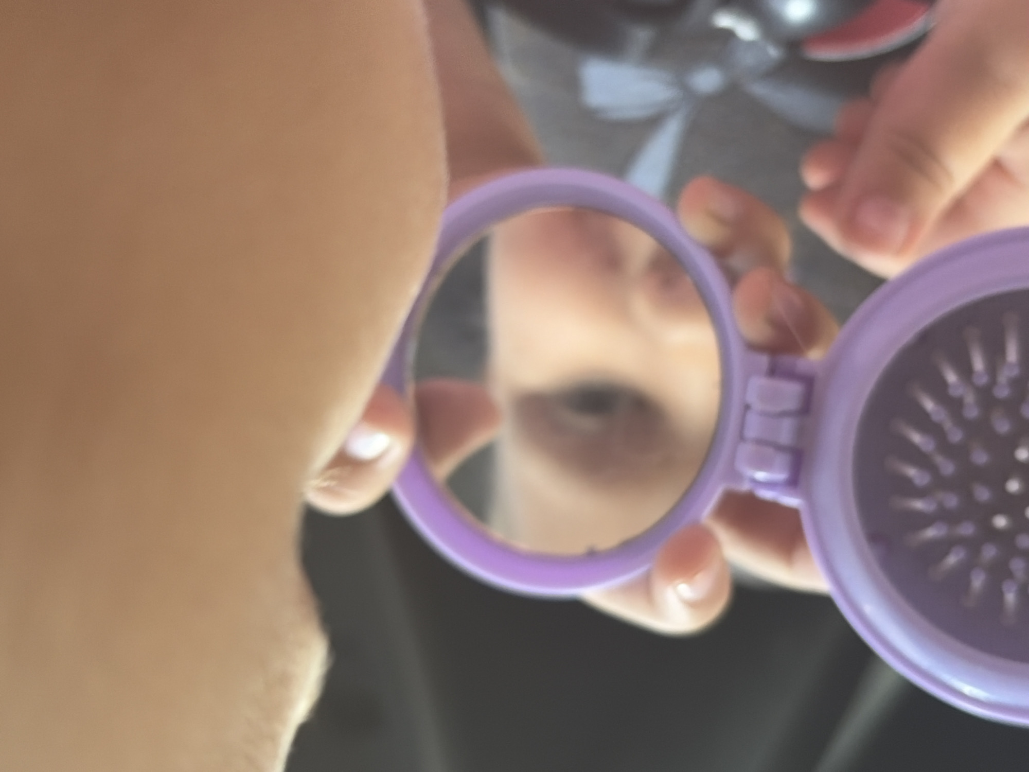 In this close-up image, a person's face is partially visible, with their nose and mouth in focus. They are holding a small, purple, oval-shaped compact mirror with a brush attached to one side. The mirror reflects the person's nose and possibly their eye, which appears to be looking into the mirror. The background is blurred, but suggests an indoor setting with dark surfaces. The image does not contain information about the location in Rostov-on-Don, Russia.
