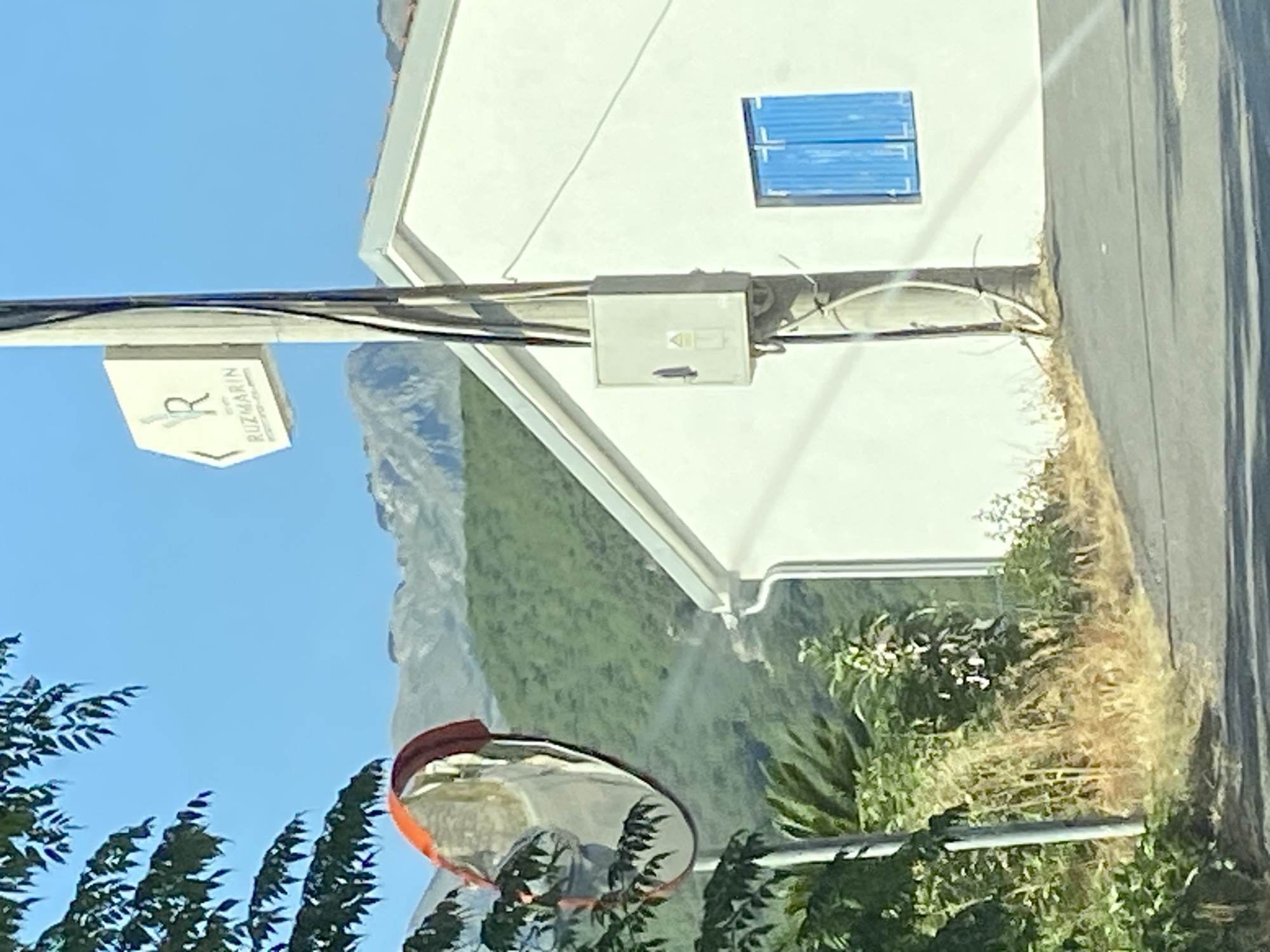 A convex mirror is positioned on the left side of the image, mounted on a metal pole that extends from the bottom of the frame upwards. This mirror reflects a road and some greenery. To the right of the mirror and pole, a white building with a white roof is visible. The building has a single window with blue shutters. Behind the building and extending across the background are rolling green hills and mountains under a clear blue sky. A sign with a "R" logo and the word "RUZMARIN" is attached to the pole, indicating a location in Seoce, Montenegro. The scene appears to be a roadside view on a sunny day.