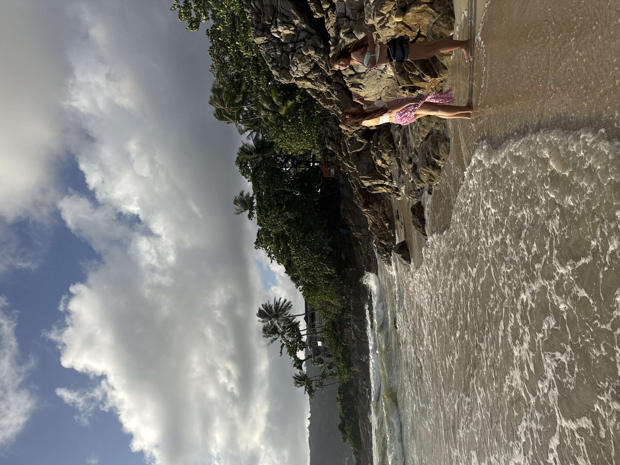 Two young women stand on a rocky beach in Patong, Thailand. The woman on the left wears a white bikini top and a pink patterned sarong. The woman on the right wears a white bikini top and dark shorts. They are standing on wet sand, with waves breaking on the shore. The beach is lined with rocks and lush green vegetation, including palm trees. In the background, a building with a dark roof is visible among the trees. The sky is cloudy, with patches of blue sky showing through. The sun appears to be low in the sky, casting long shadows. The overall atmosphere is one of a tropical, coastal setting.
