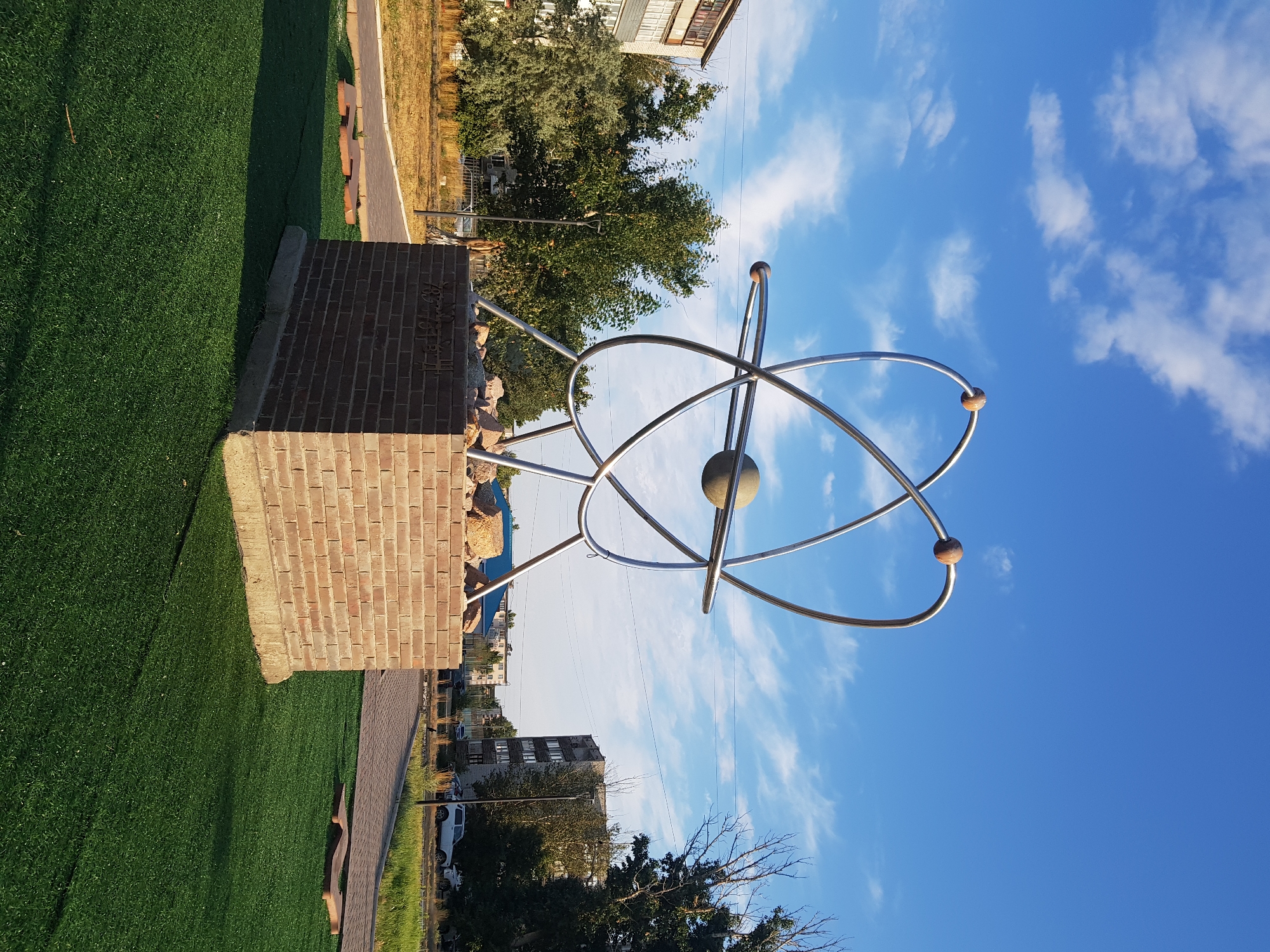 The image shows a monument in the city of Kurchatov, Kazakhstan, that symbolizes an atom with orbiting electrons. The monument is made of polished metal and is mounted on a brick pedestal. The pedestal is surrounded by artificial turf. In the background, there are buildings, trees, and a blue sky with clouds. The monument appears to be located in a public space, possibly a park or a plaza.