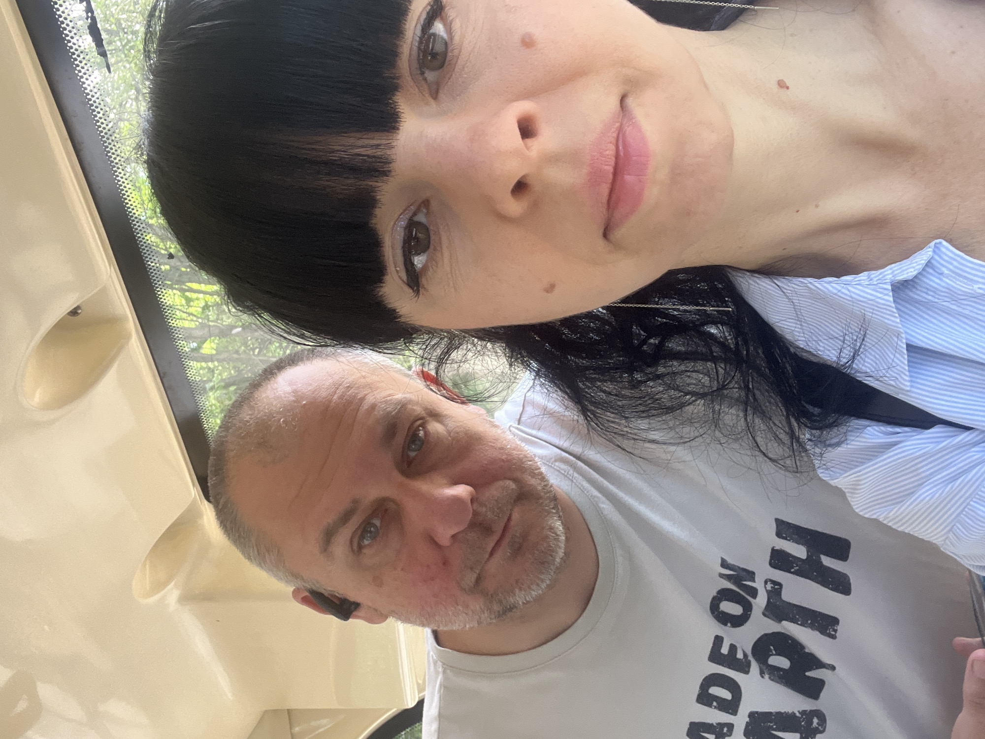 Two people are posing for a selfie. A woman with dark hair and bangs is on top, with her face close to the camera. She is wearing a light blue striped shirt. Below her is a man with short, graying hair and a beard. He is wearing a white T-shirt with large, black lettering that reads "MADE ON EARTH". A black object is visible in his left ear. They are inside a vehicle with a light-colored interior. Through the windshield, trees and greenery are visible, suggesting an outdoor setting. It is not possible to determine if this photo was taken in the city of Krasnodar, Russia.