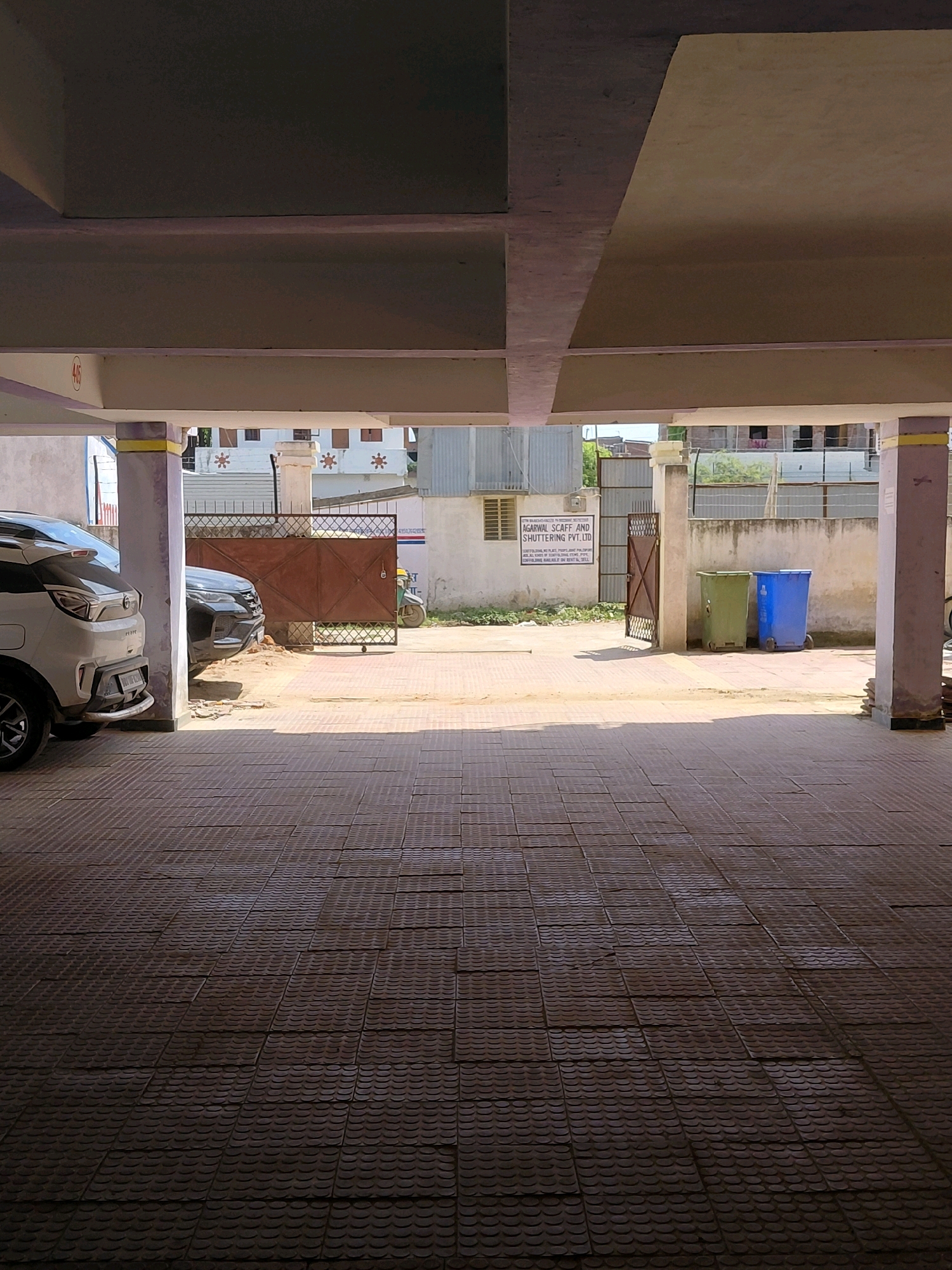 The image shows a covered parking area with tiled flooring. Two cars are parked within the area, one white and the other black. Beyond the parking area, a metal gate is visible, leading to an outdoor space. A green and a blue trash bin are placed outside the gate, to the right. A sign with text in English and Hindi is mounted on the wall near the gate, appearing to be an advertisement or official notice for "AGARWAL SCAFF AND SHUTTERING PVT. LTD." A motorcycle is parked outside the gate. The background includes buildings with distinct architectural features, suggesting an urban setting in Patna, India. The overall lighting indicates it is daytime, with shadows cast across the tiled floor, implying sunlight is entering from the side. The environment appears to be a residential or commercial complex.